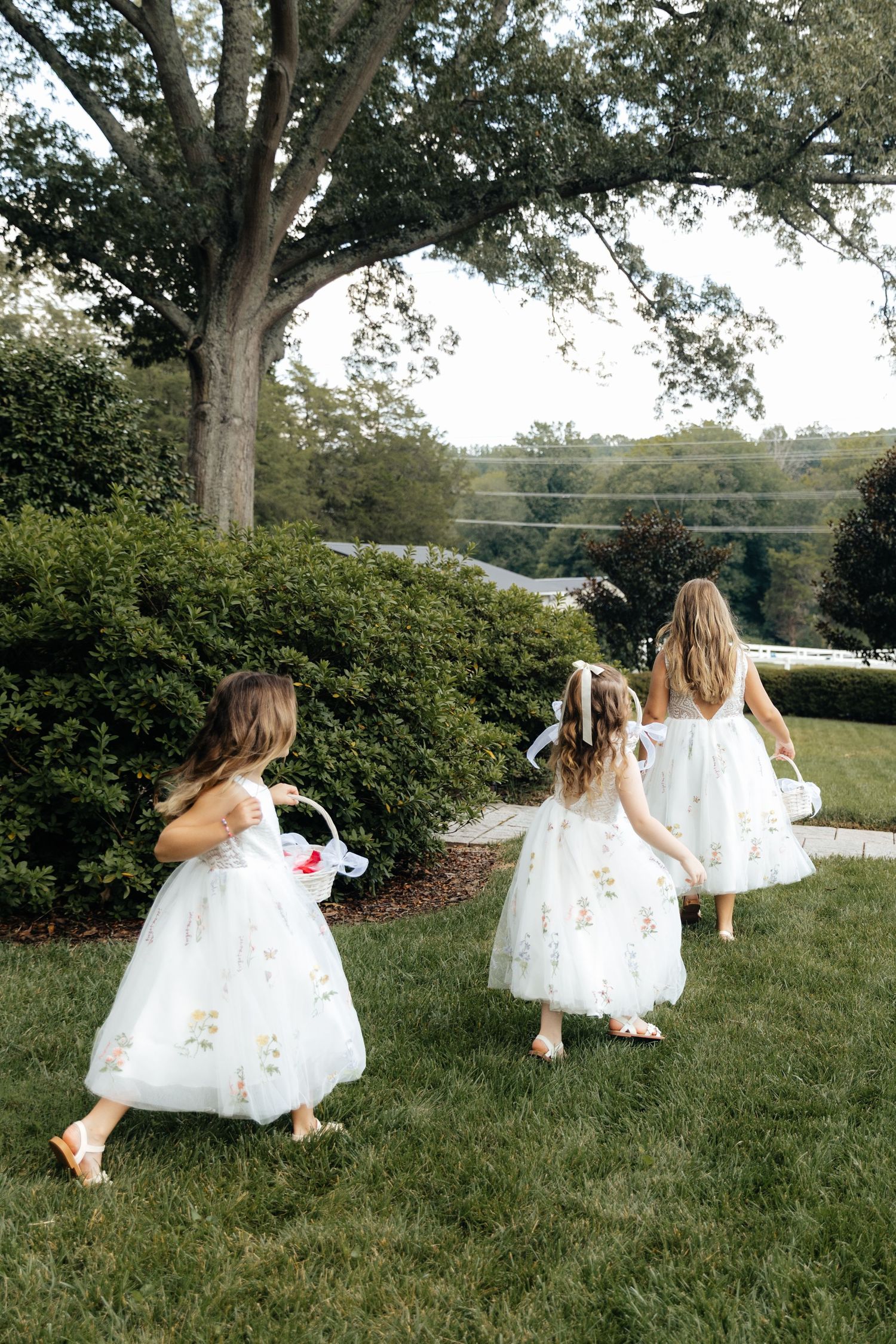 Flower girls in white dresses running and playing on a grassy lawn near large trees.