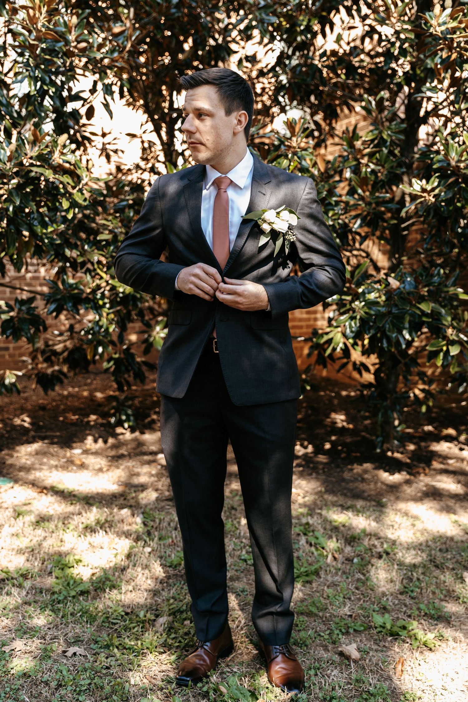 Groomsmen in dark suits adjusting their ties outdoors near foliage.
