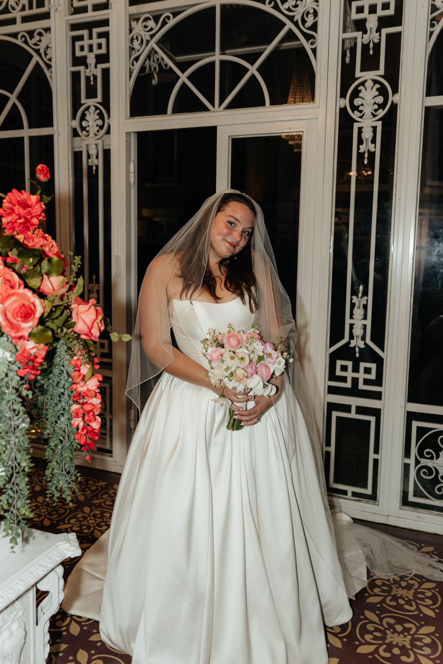 Bride poses with pink bouquet against decorative iron and glass conservatory backdrop.