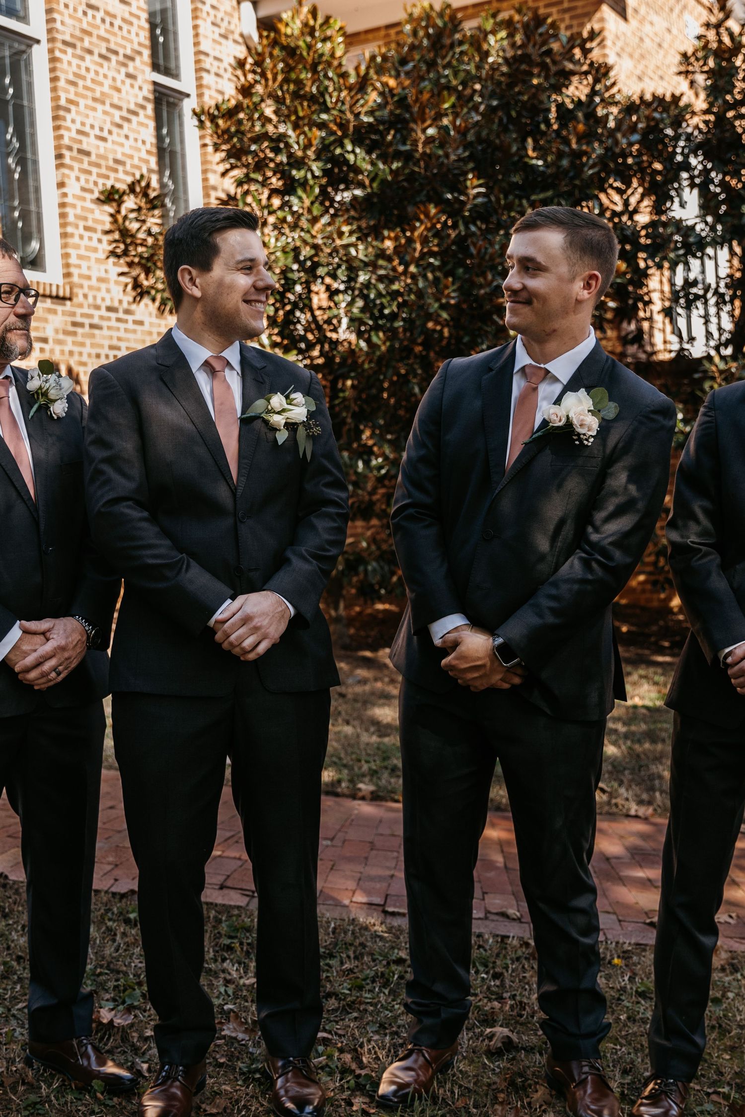 The groom and groomsmen in dark suits with coral ties share a moment outdoors.