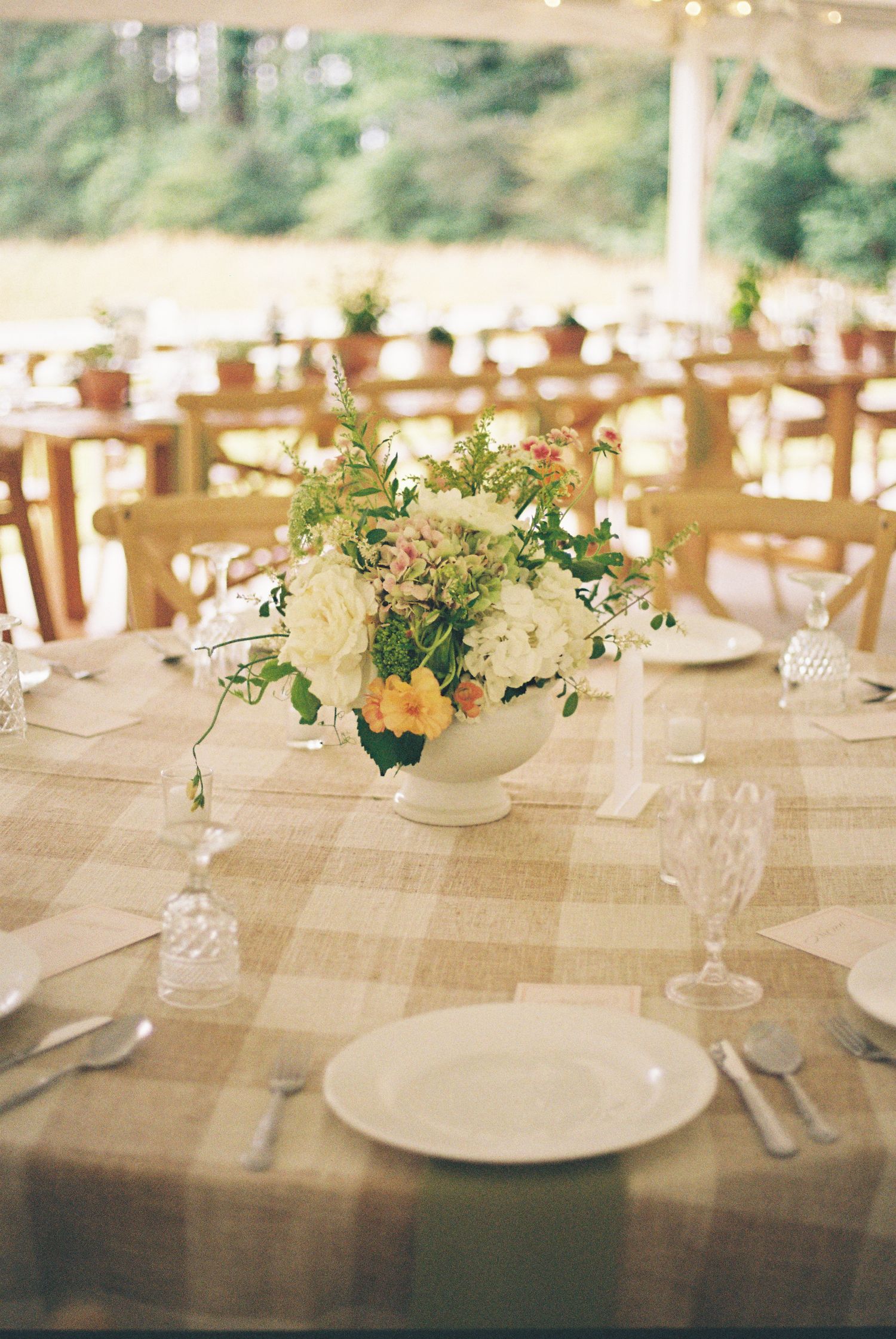 Elegant wedding reception table with plaid linen, white floral centerpiece, crystal glasses, and white plates under a tent.