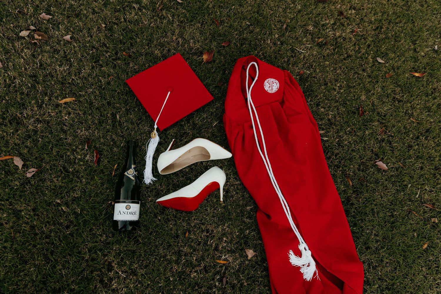 Red graduation cap, gown, white heels, and champagne bottle arranged on green grass for a celebratory graduation photo.