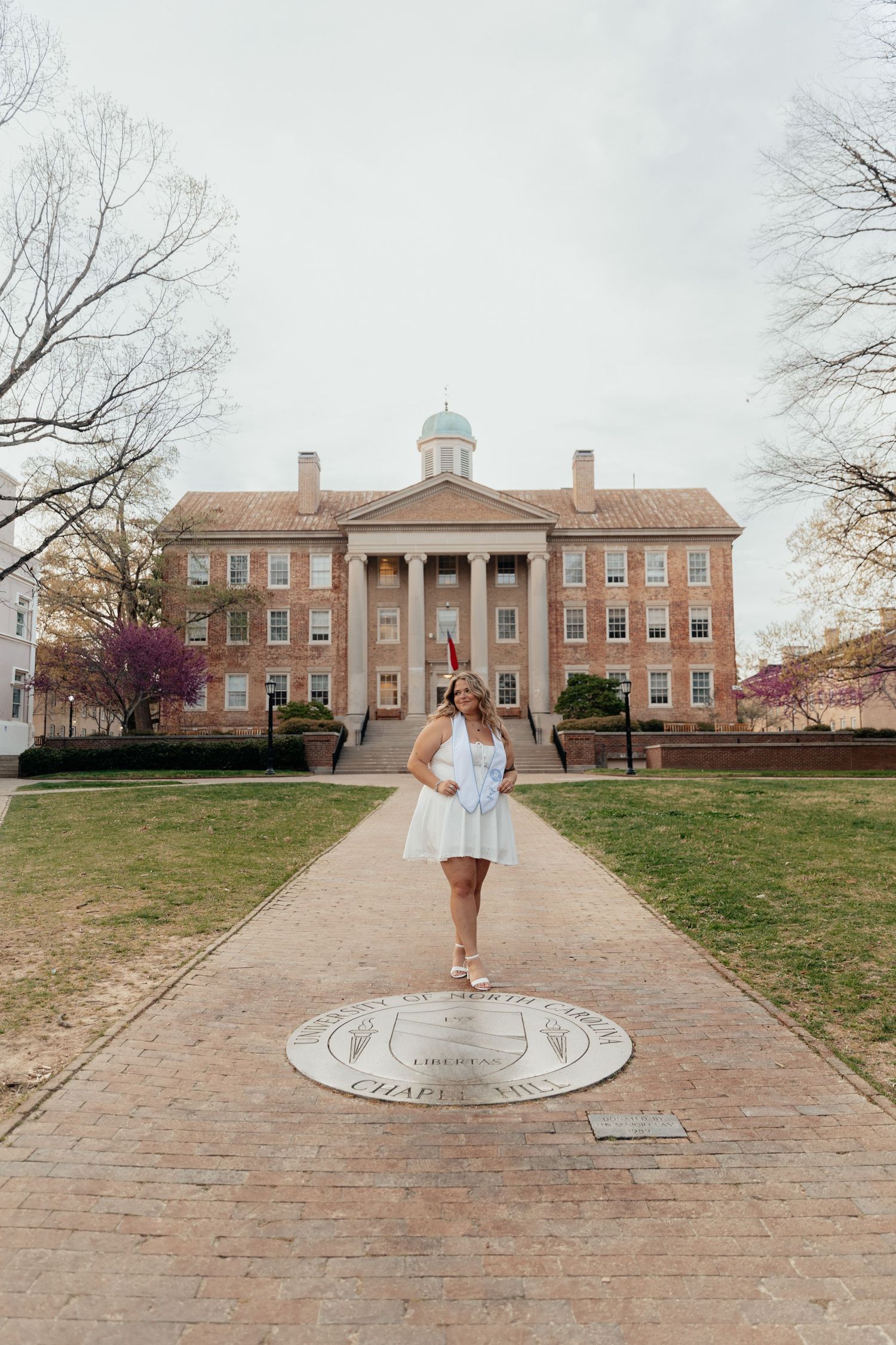 College graduate in white dress stands on UNC Chapel Hill seal outside historic South Building on campus.