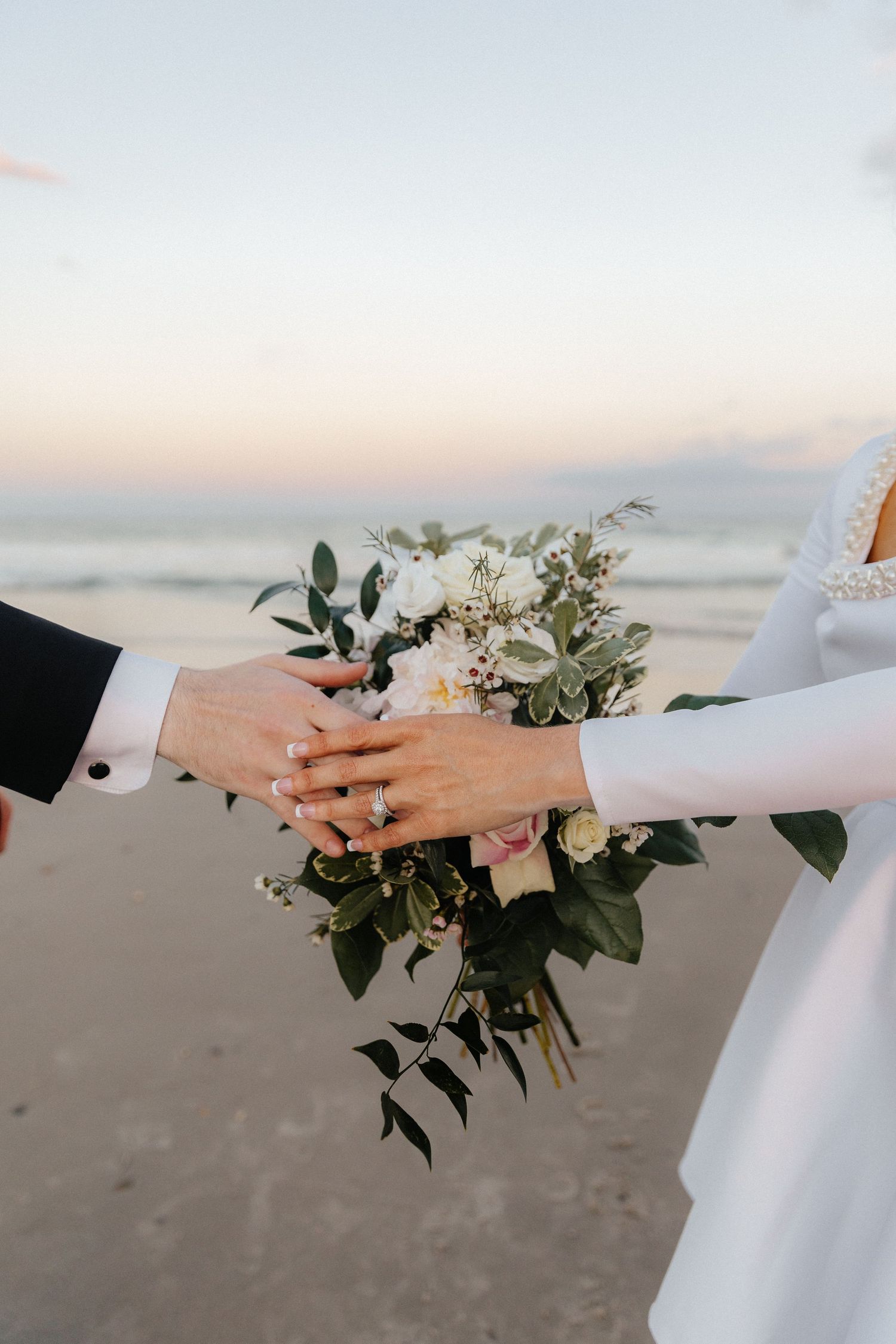 Wedding bouquet detail shot with delicate white flowers and greenery against beach backdrop.