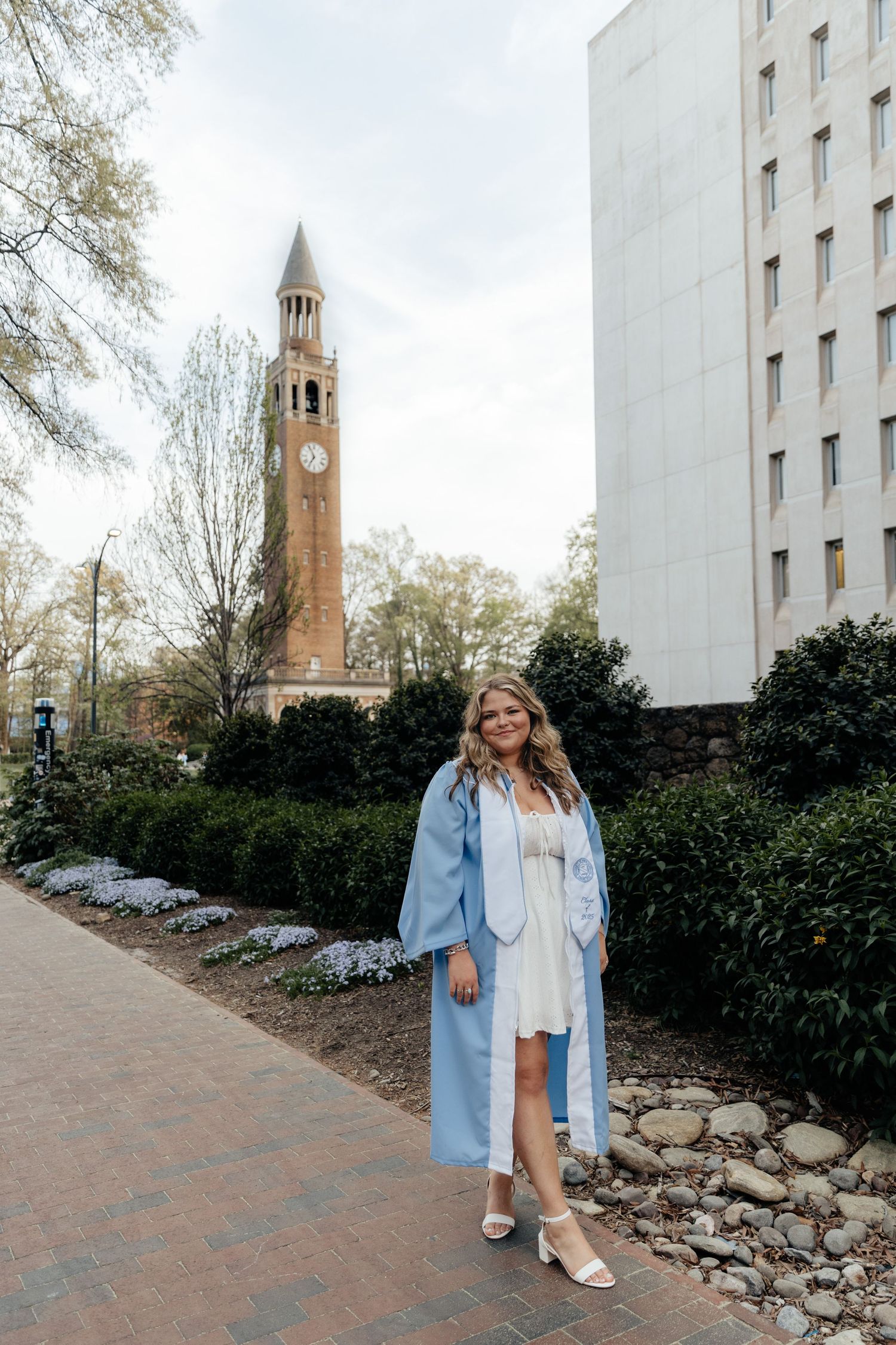 Graduate in light blue regalia poses near historic clock tower on campus.