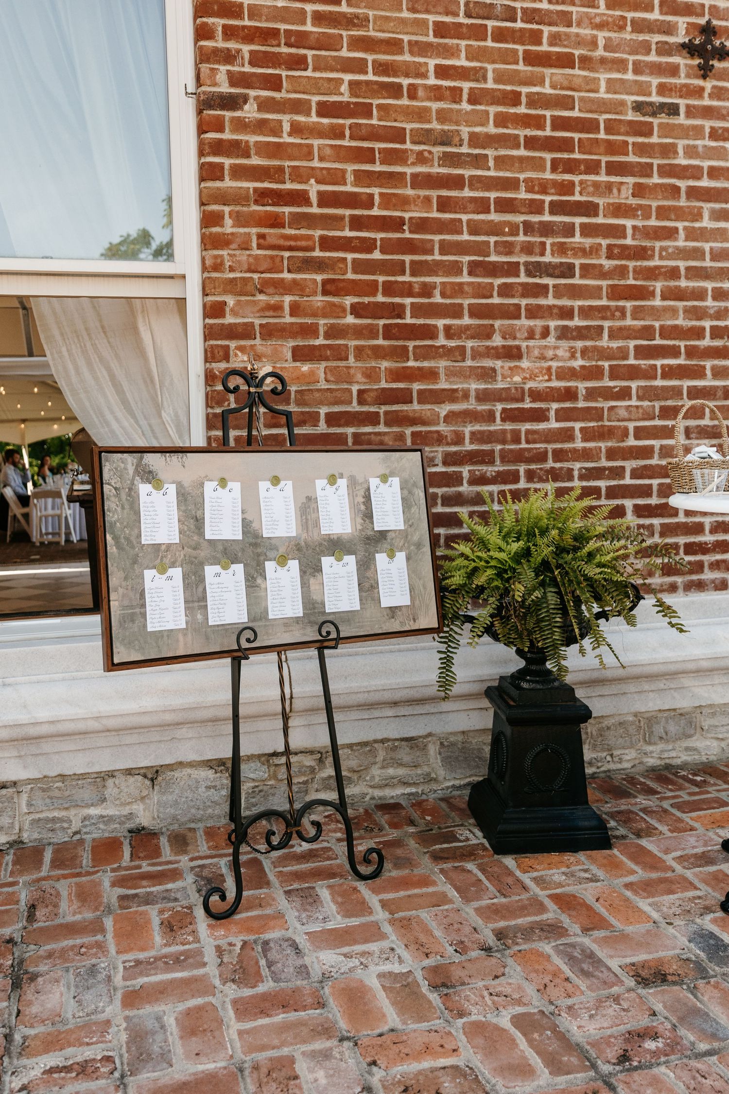 Wedding seating chart display on metal stands against brick wall with potted plants.