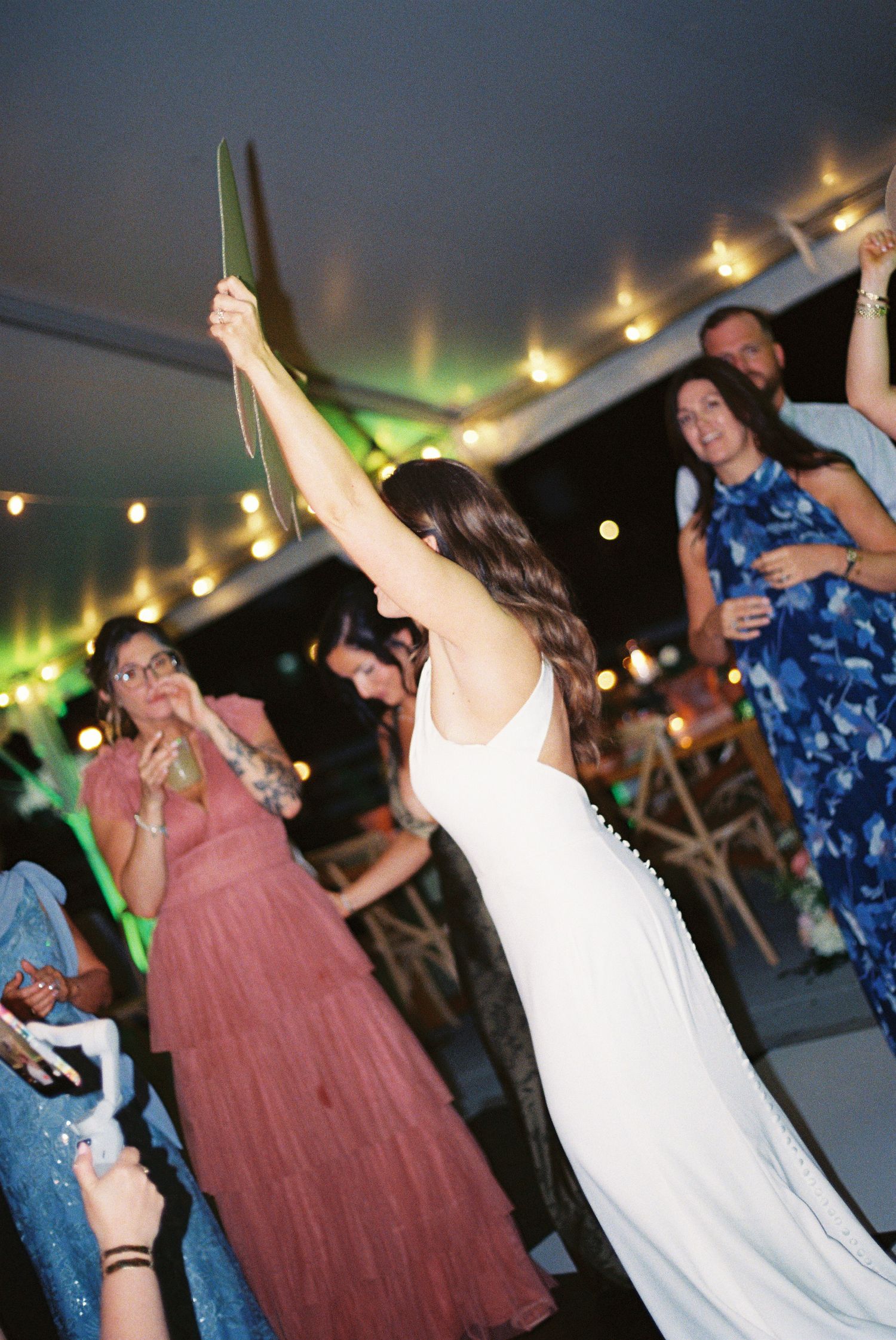 Bride in white gown raises arm holding greenery on wedding reception dance floor with guests celebrating.