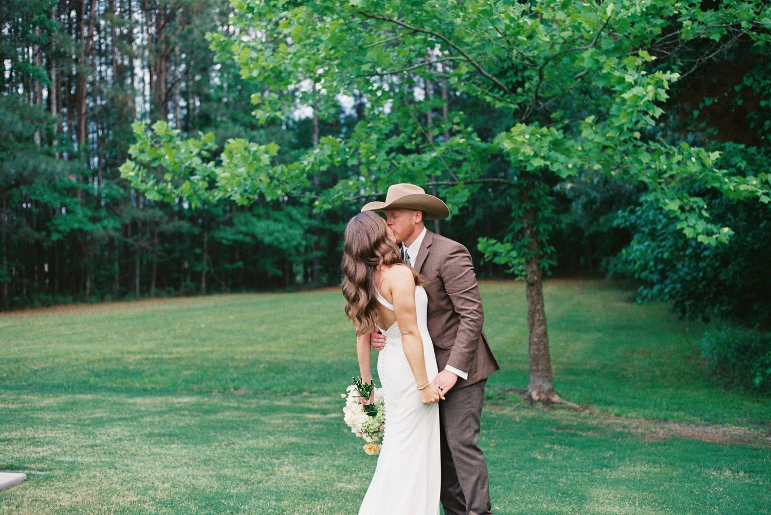 Bride and groom sharing a romantic kiss in a lush green outdoor setting, groom wearing a brown suit and cowboy hat.