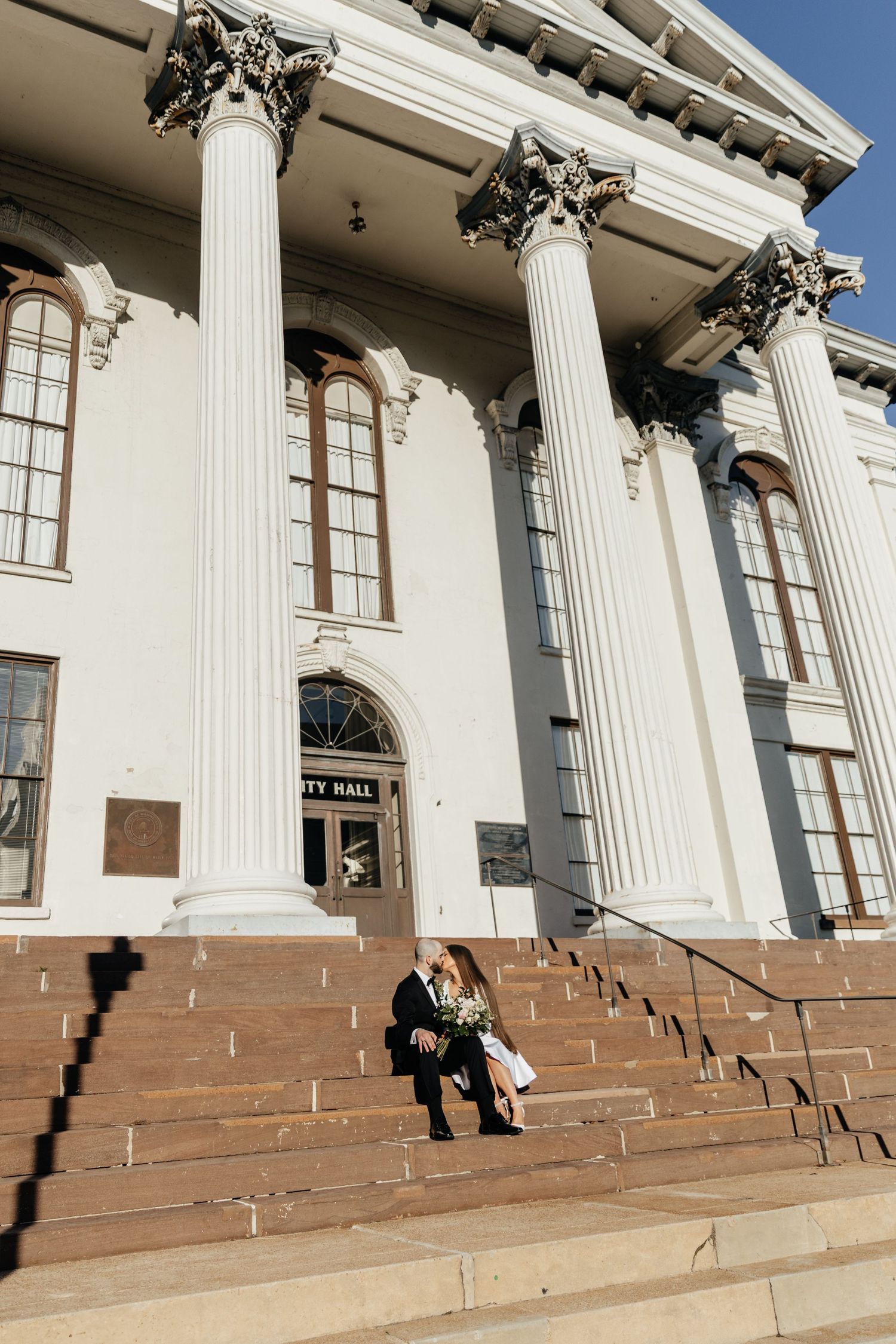 Bride and groom sitting on the steps of a white building facade with tall columns.