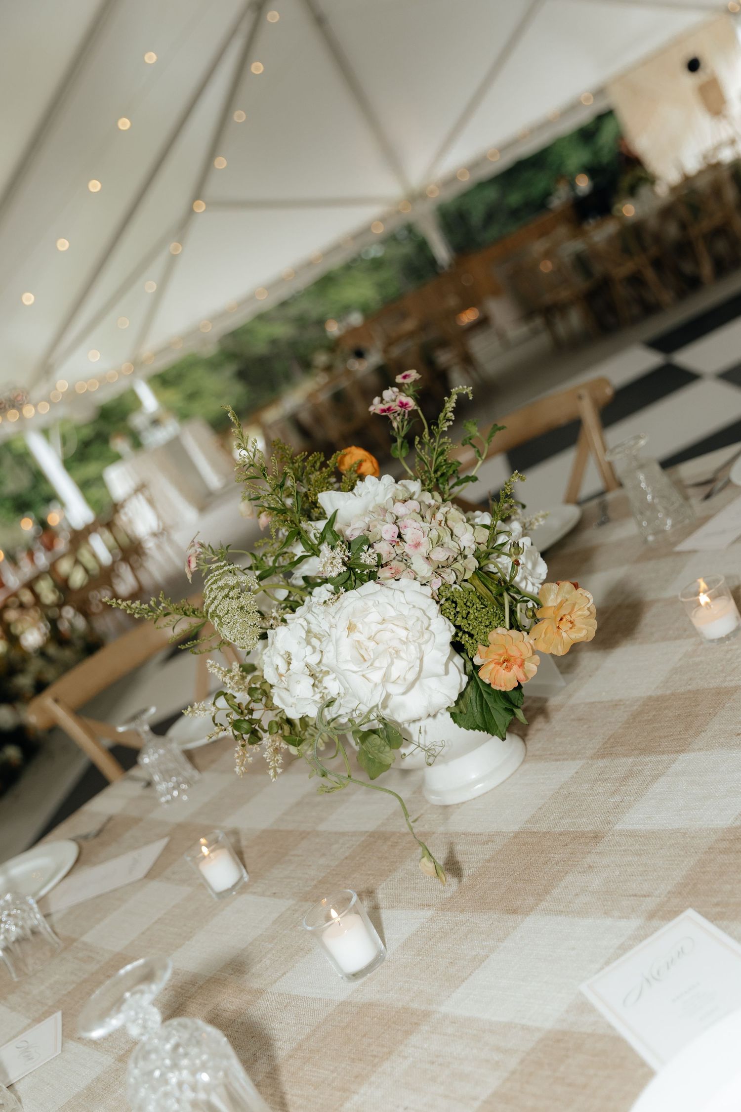 Elegant wedding reception centerpiece with white peonies, peach roses, and greenery on a gingham tablecloth under a tent.