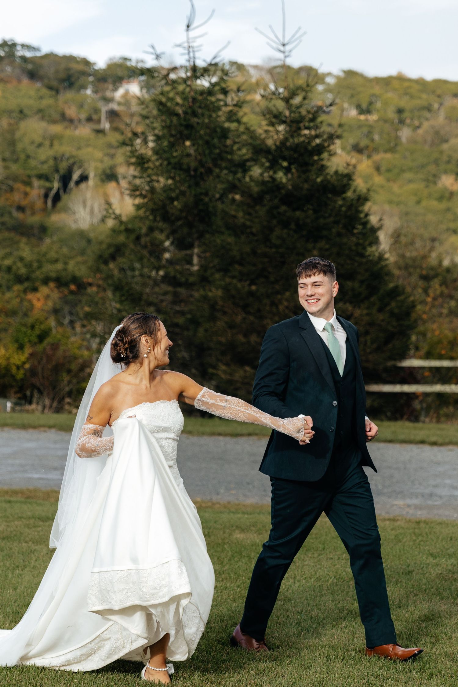 Bride in white gown and groom in navy suit walk hand in hand on green lawn with wooded hills backdrop.