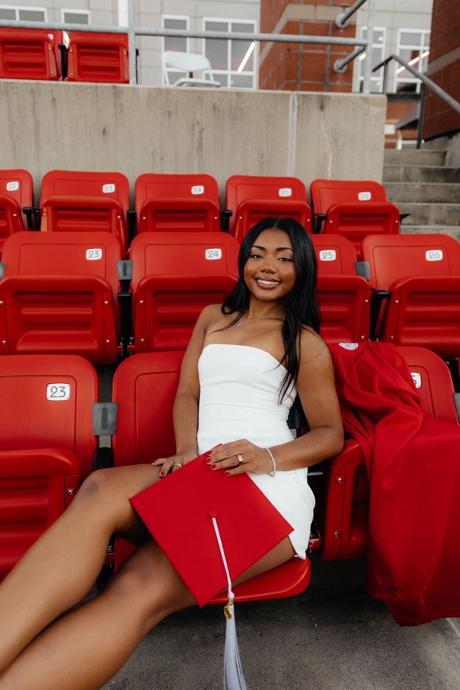 Smiling graduate in white dress holding red cap seated in stadium bleachers.