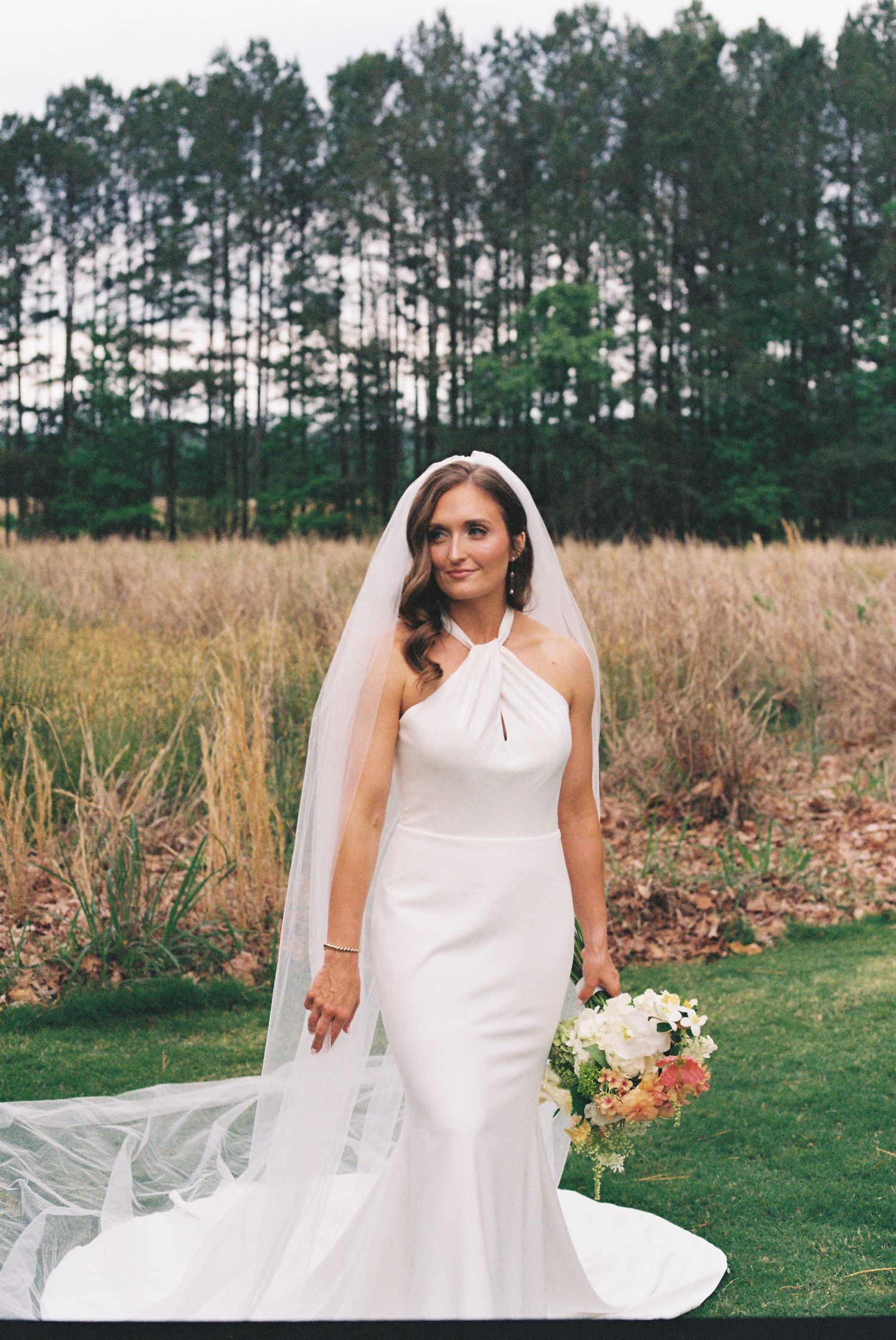 Bride in white halter mermaid gown and veil holds bouquet outdoors near tall grasses and pine trees.
