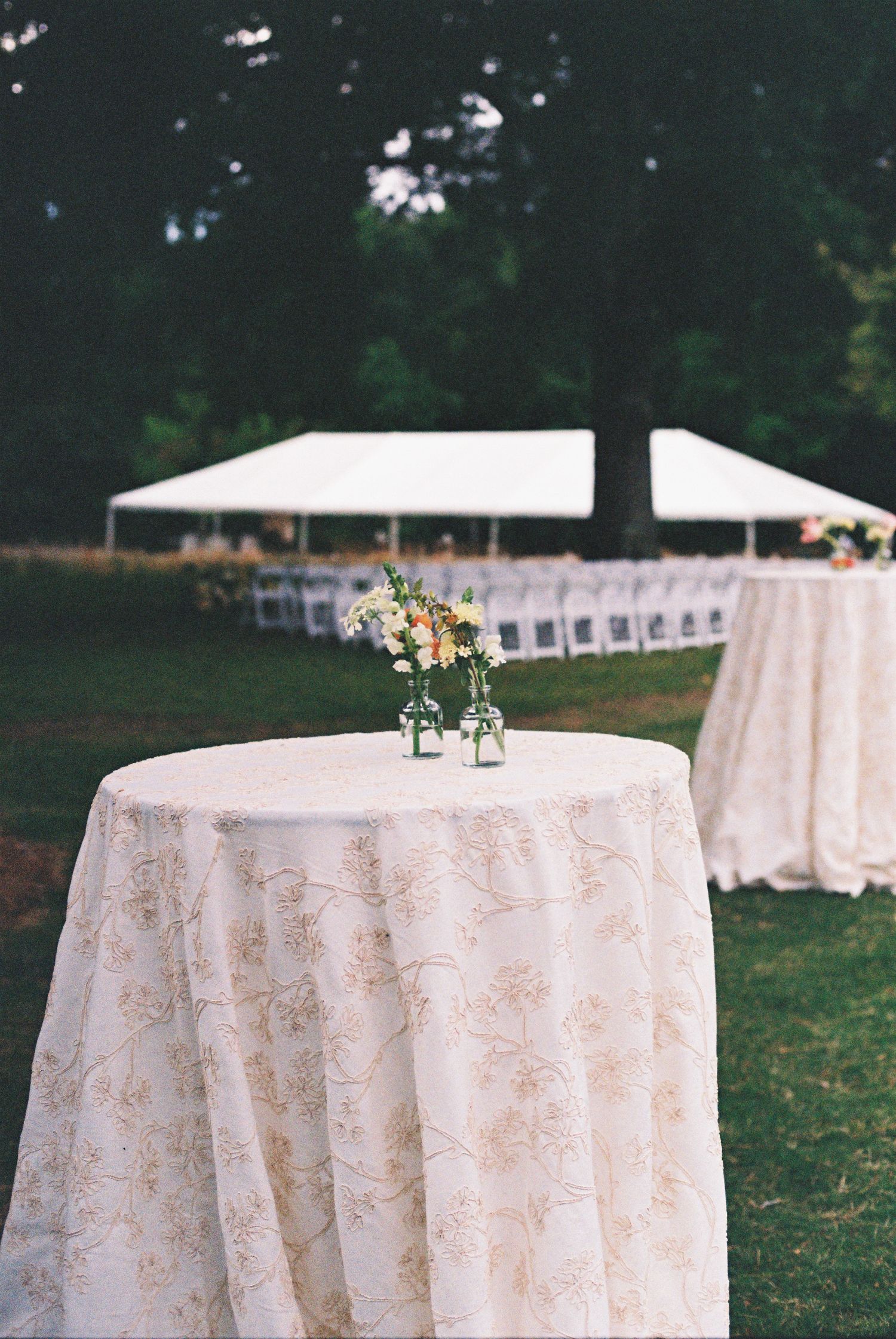 Elegant outdoor wedding reception with floral tablecloths, small flower centerpieces, and a white tent with chairs in the background.