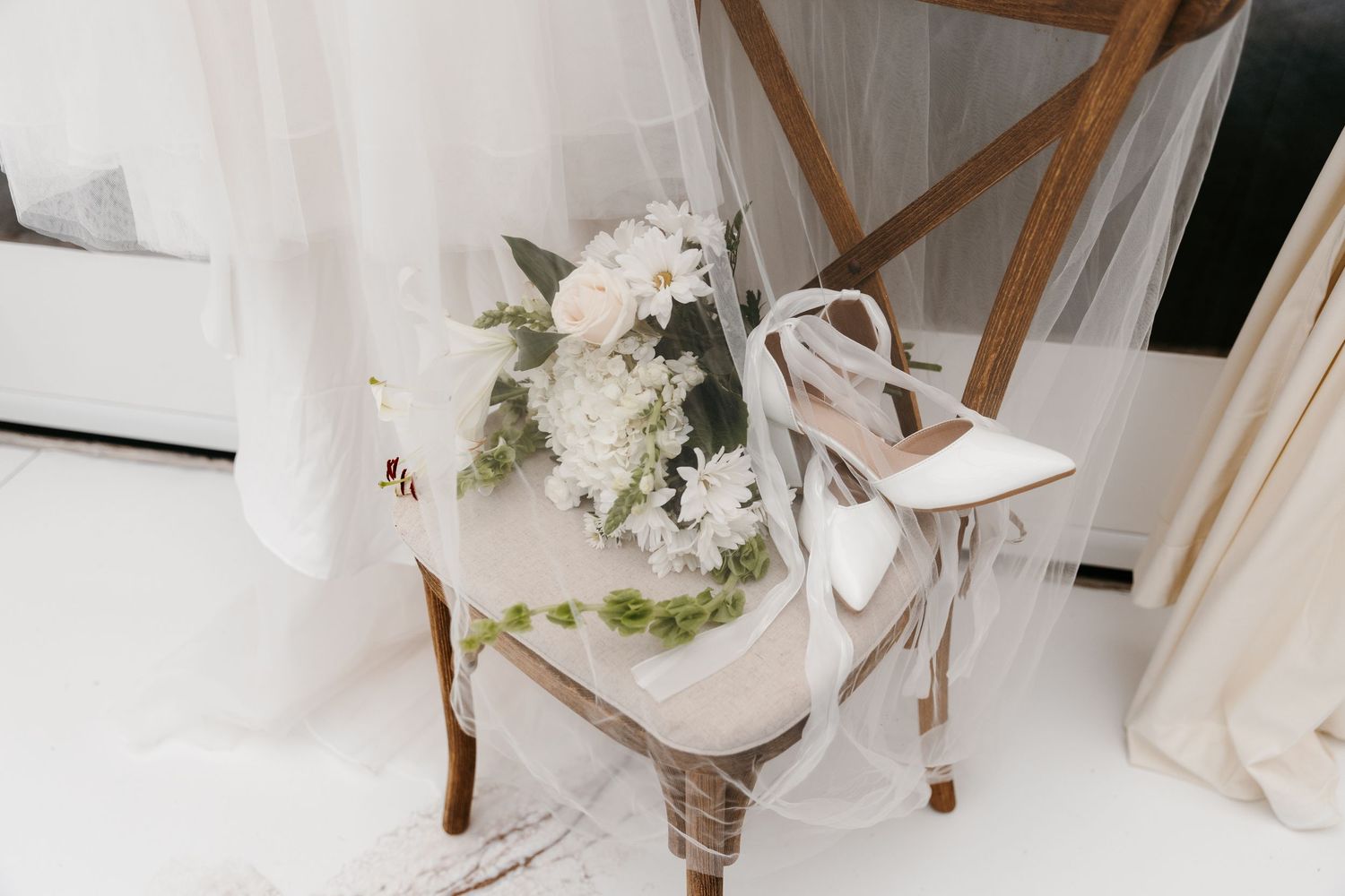 Elegant bridal setup with white heels, floral bouquet, and flowing veil on a vintage chair.