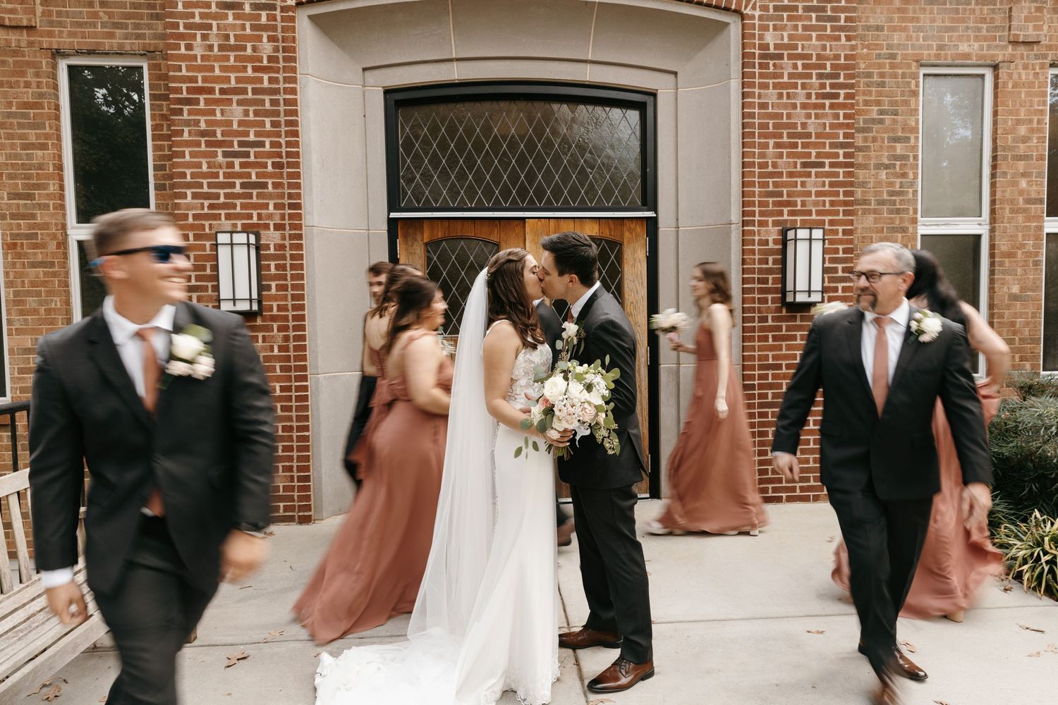 Wedding party motion-blur posing in front of brick church with couple sharing a kiss in the middle.