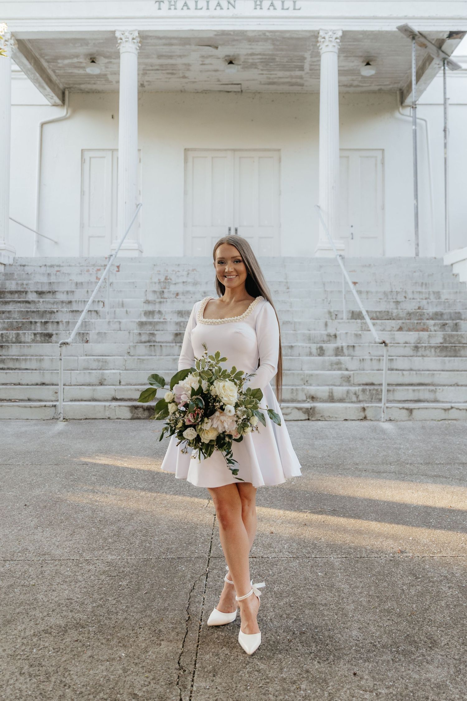 A bride holds a romantic floral bouquet while posing in a short white dress against a white building.