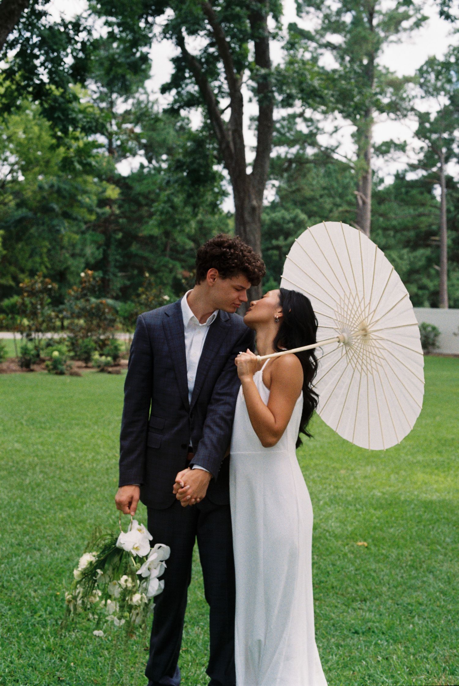 Wedding couple poses playfully with white parasol in outdoor garden setting.