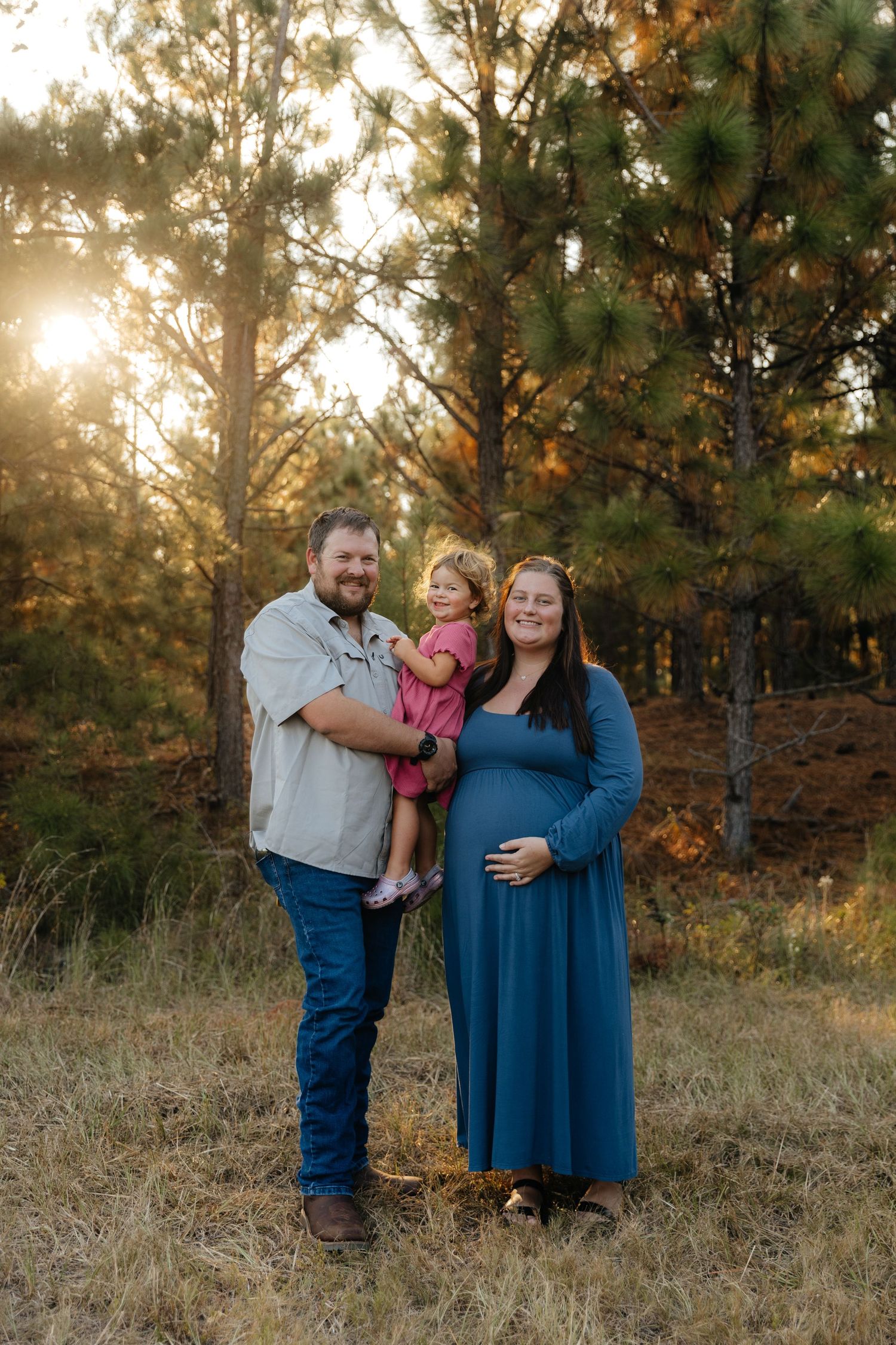 Family standing together in a pine forest during golden hour sunset.