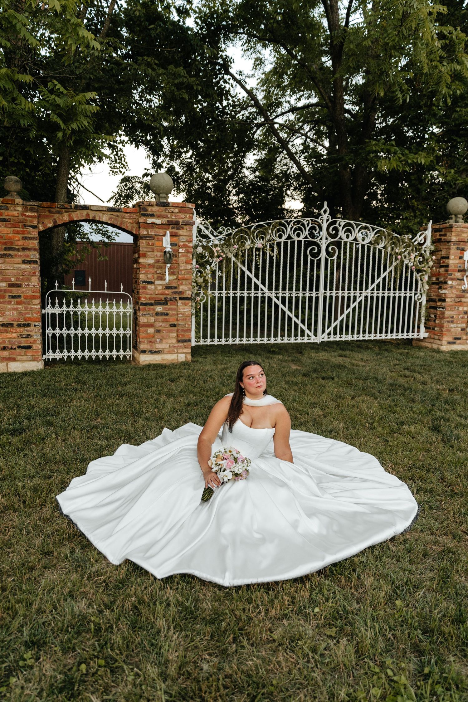 Bride in flowing white gown poses gracefully on grass near iron gate entrance.