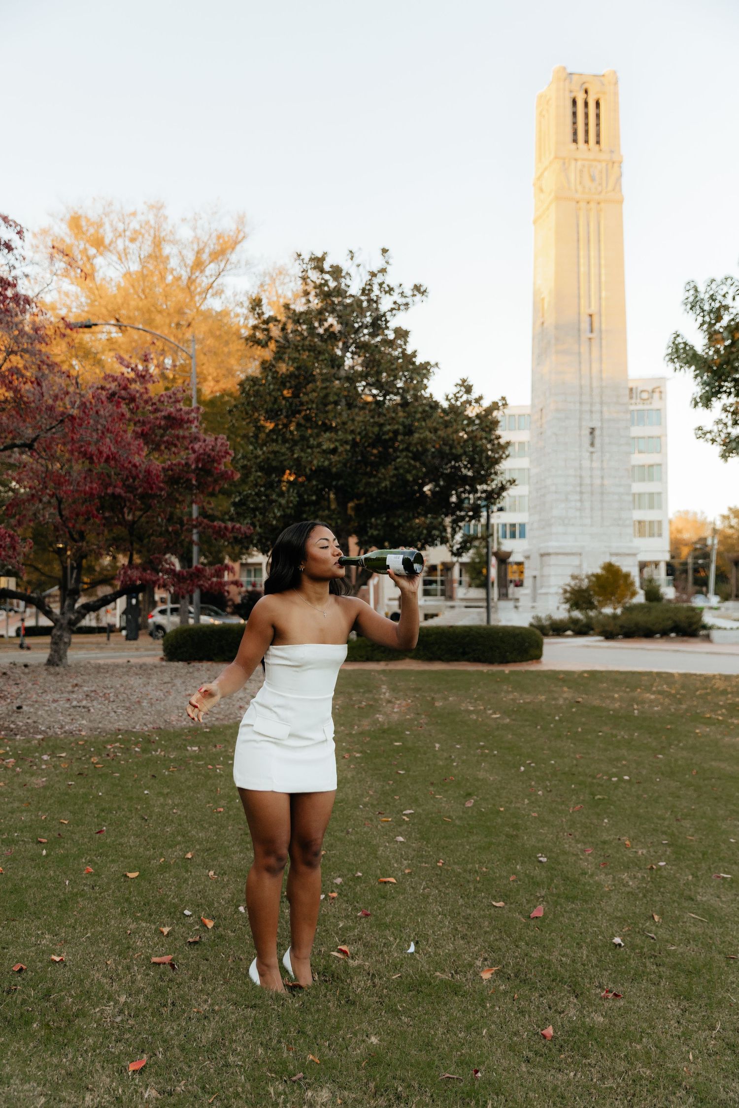 Young Black woman in white strapless mini dress poses outdoors near a tall tower and colorful autumn trees at sunset.