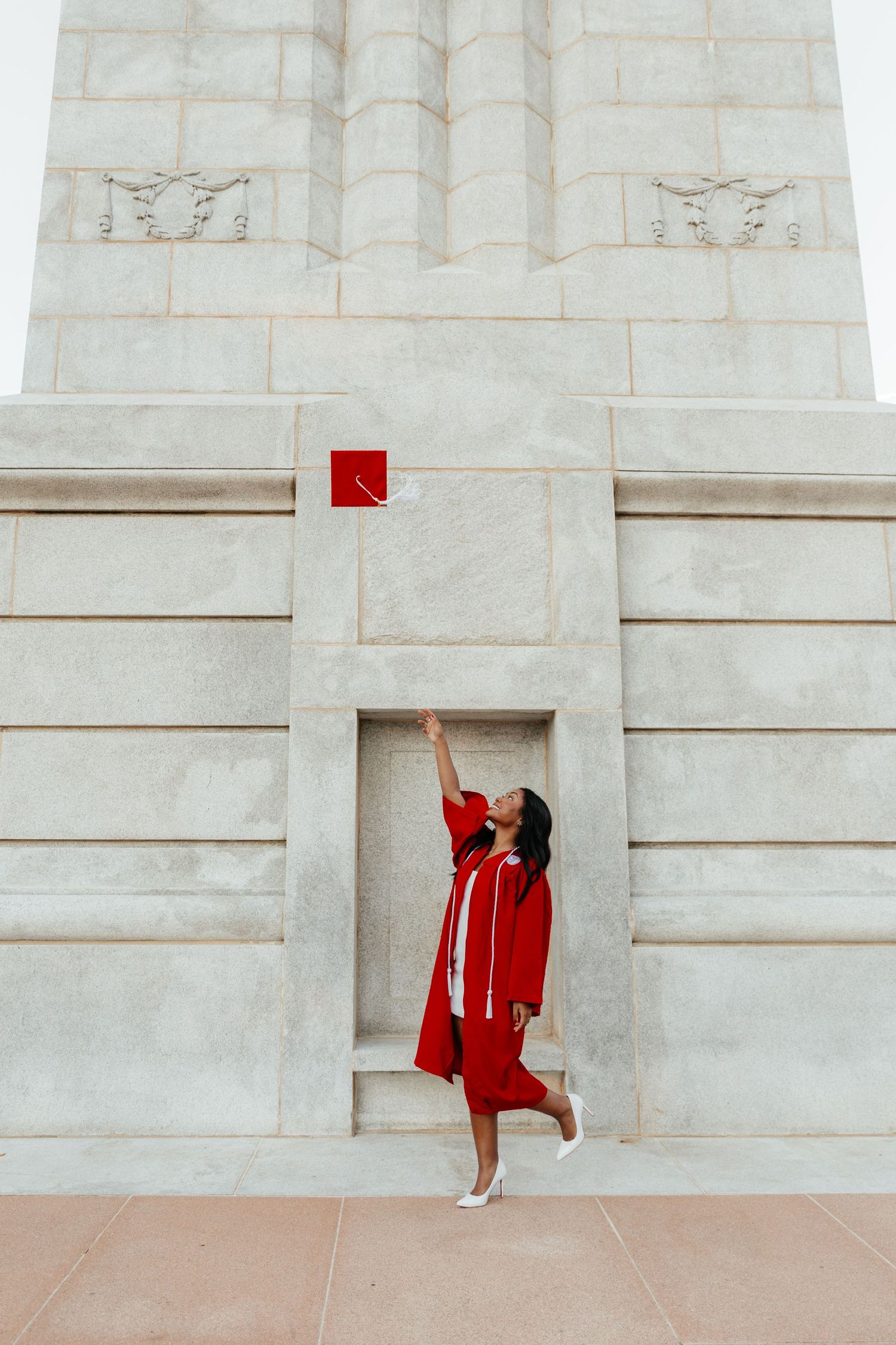 A graduate in red cap and gown tosses her mortarboard at a grand white stone monument.