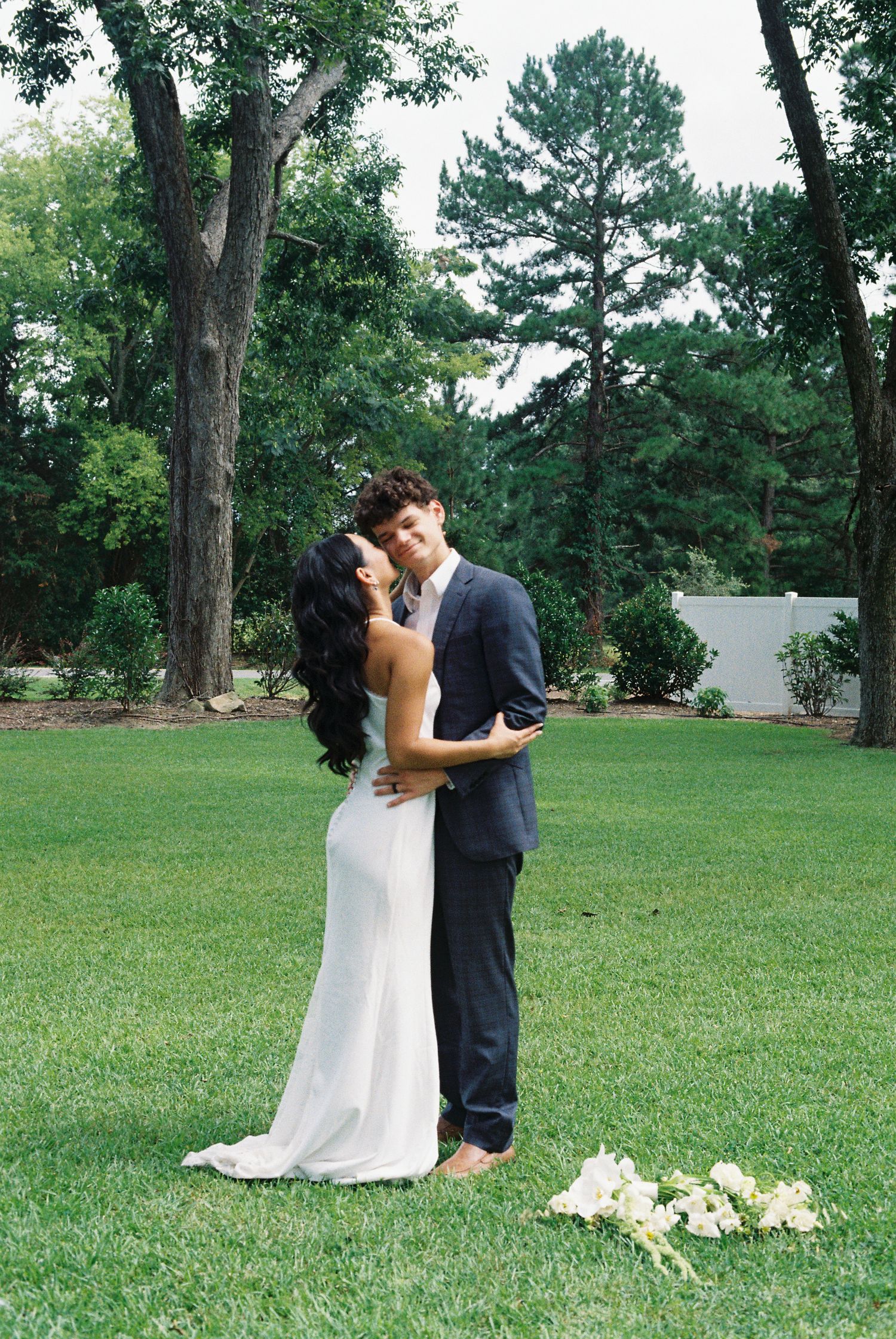Romantic wedding couple embraces while dancing on a green lawn surrounded by trees.