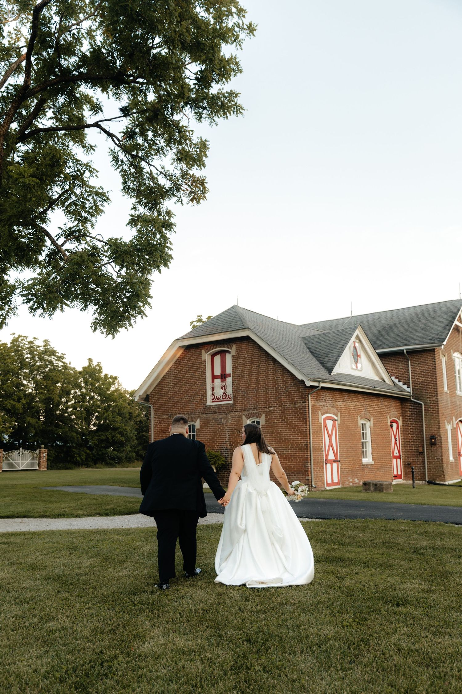 A couple in formal wedding attire walks on a grassy hill near a brick church at sunset.