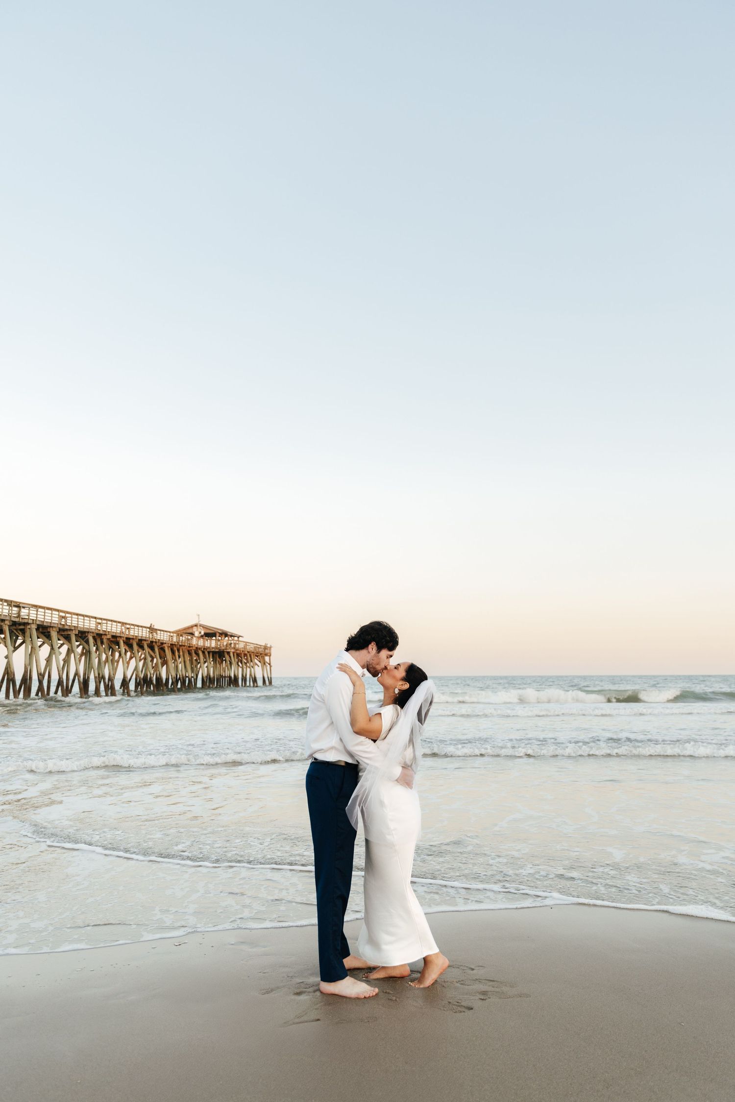 Couple sharing a romantic kiss on a beach at sunset near a wooden pier, bride in white gown and groom in navy pants.