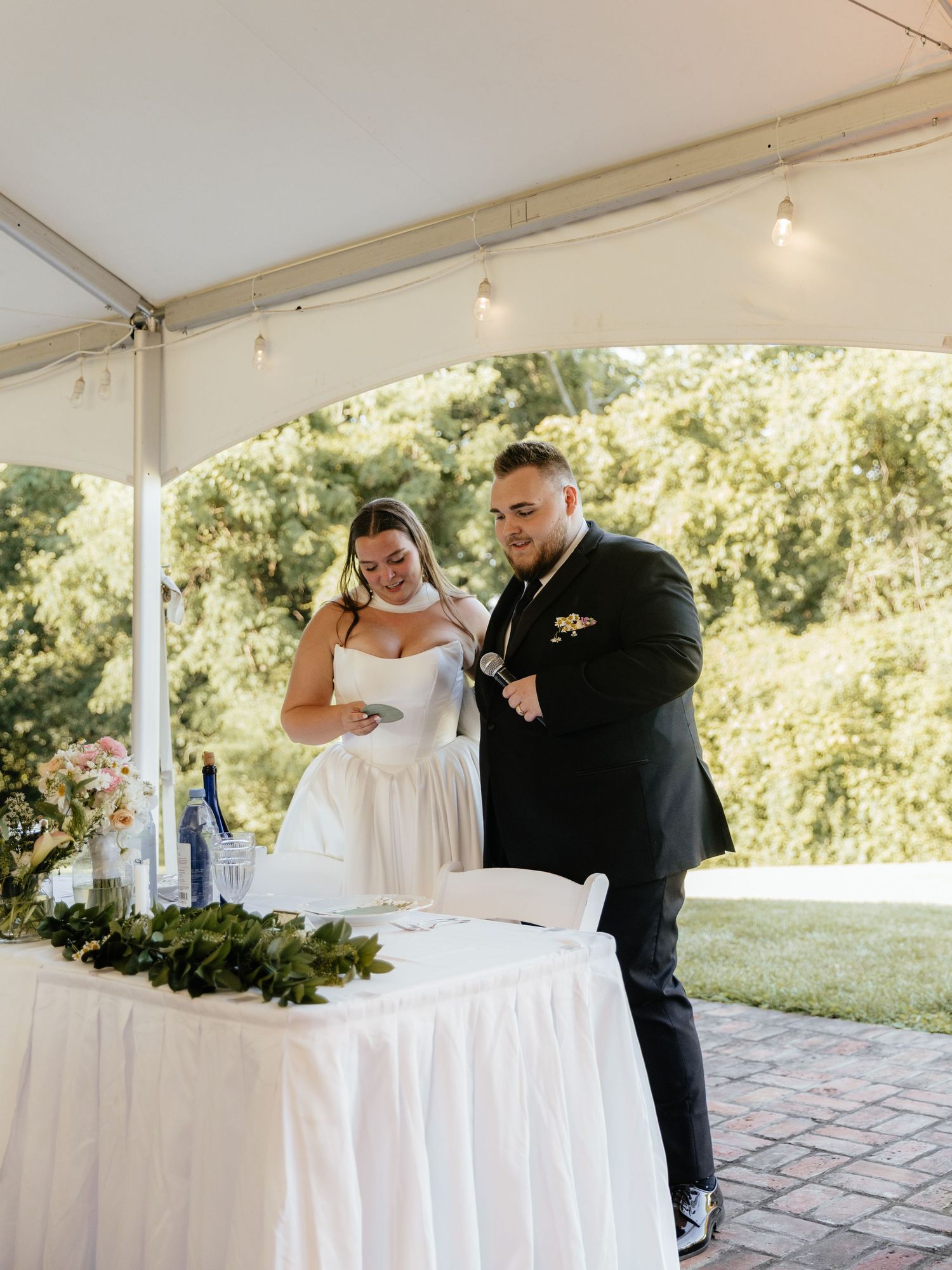 Bride and groom stand together under a white tent at an outdoor wedding reception, reviewing items at a decorated table.