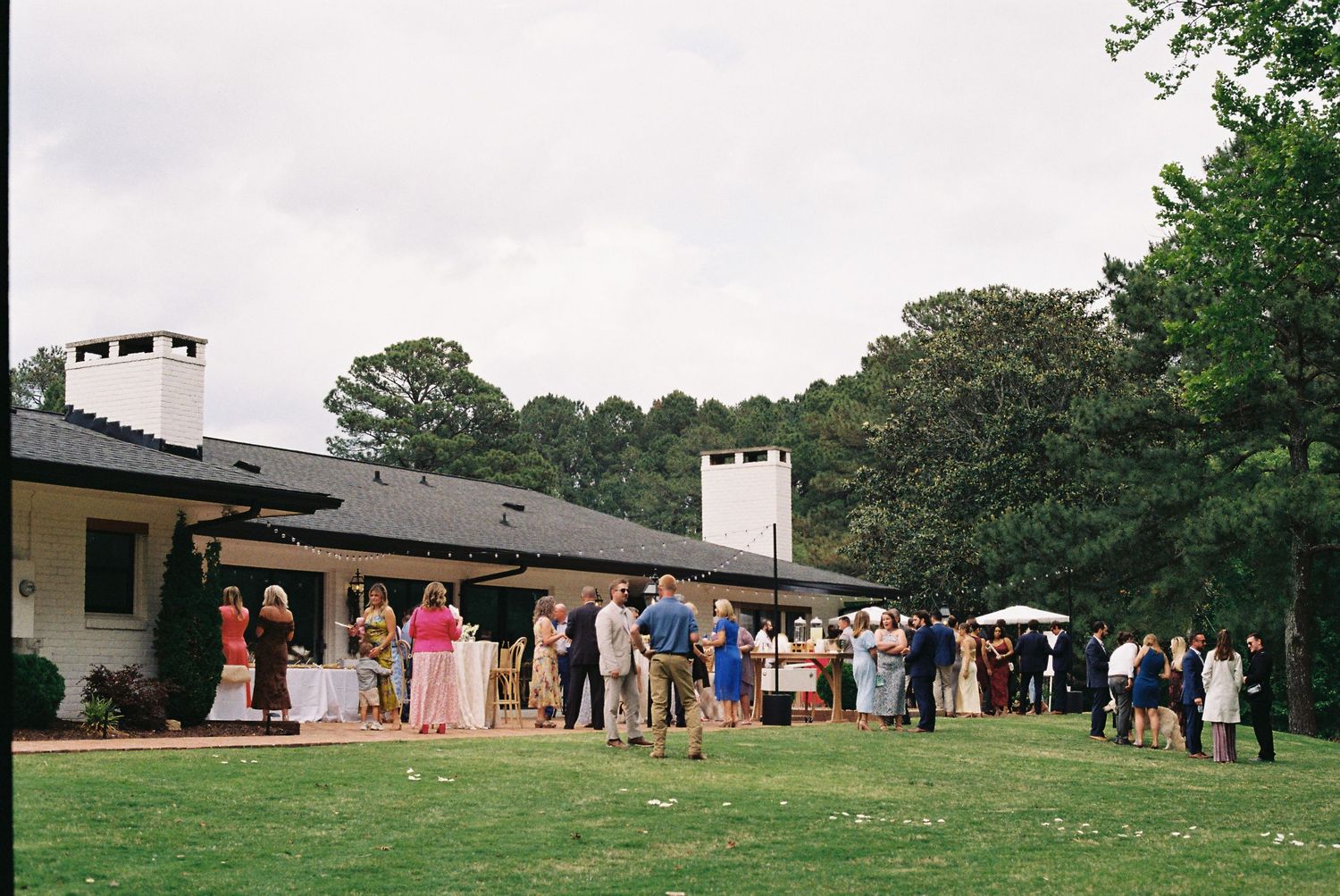 Outdoor garden party with guests mingling on a green lawn beside a low ranch-style house surrounded by tall trees.