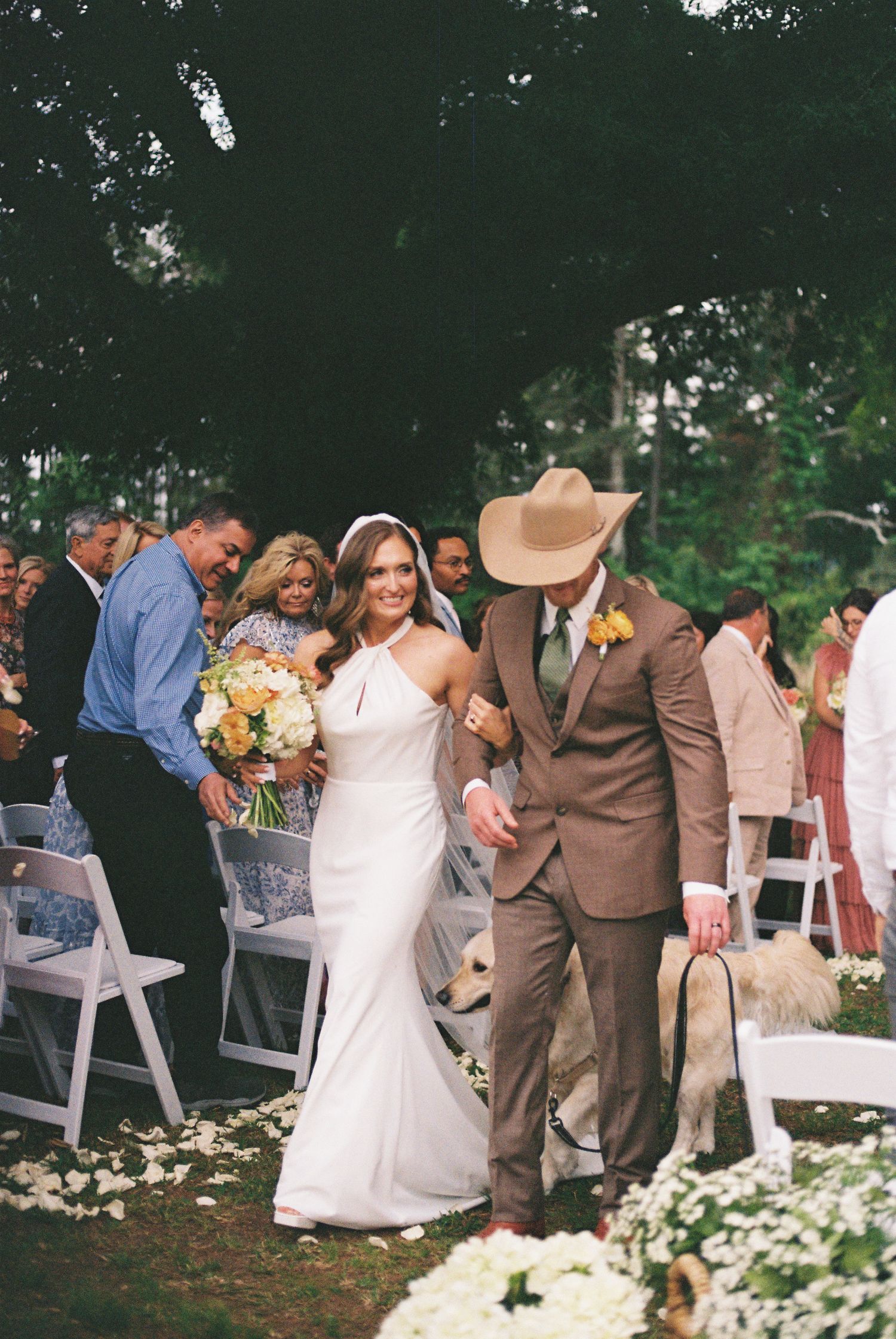 Bride and groom walk down outdoor wedding aisle with dog, groom wearing brown suit and cowboy hat.