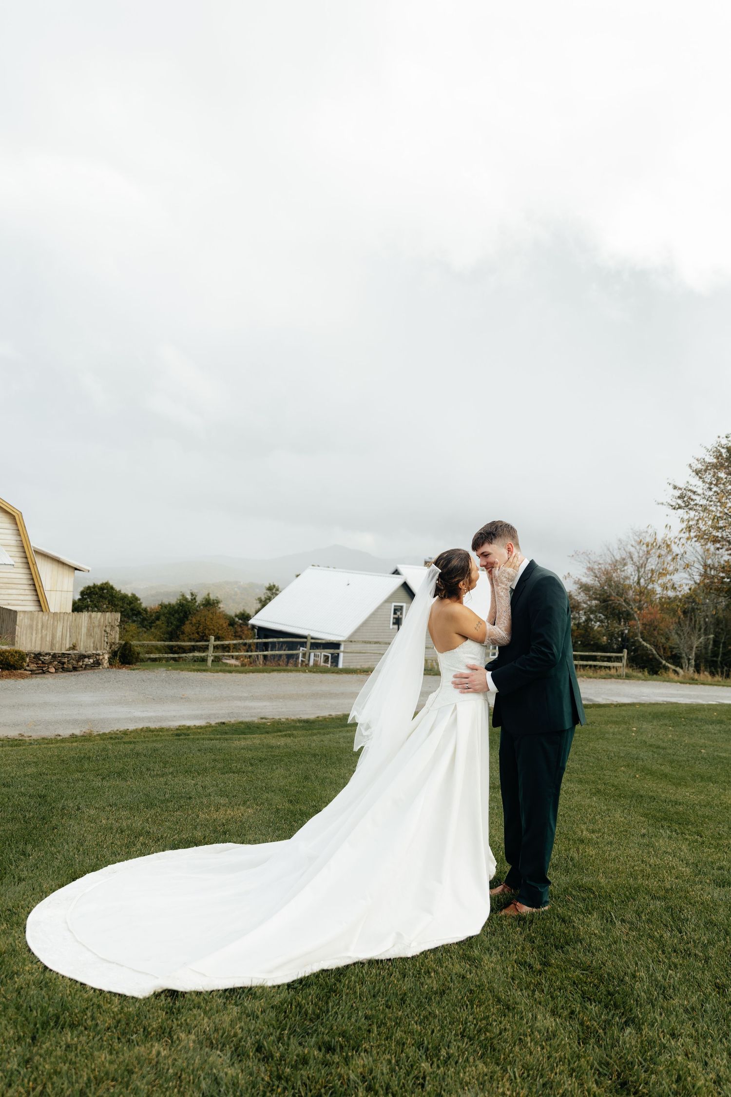A bride and groom share a romantic moment on green grass, with her white gown trailing behind them at a rustic farm venue.