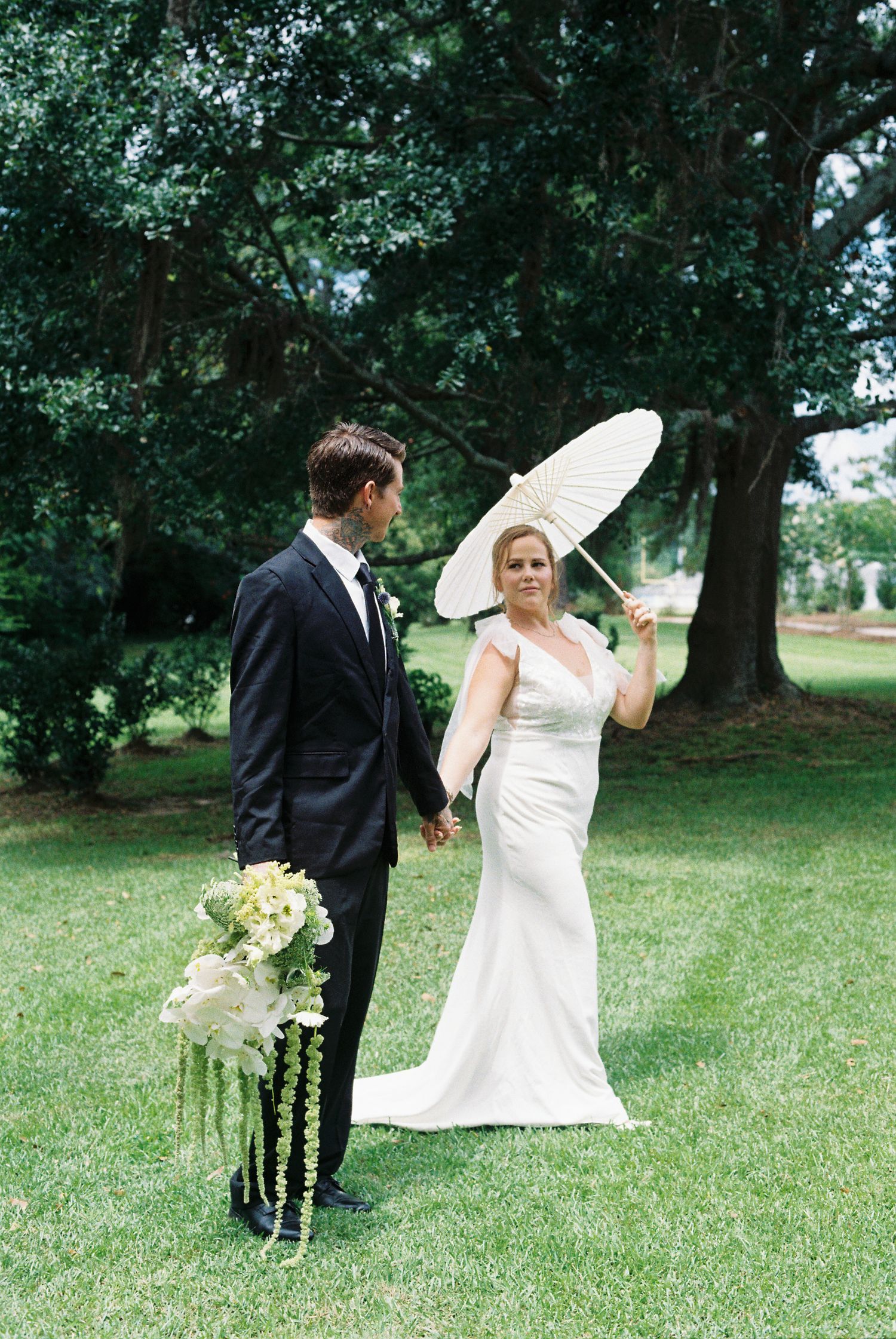 Elegant wedding photoshoot in a garden setting with white parasol and flowing dress.