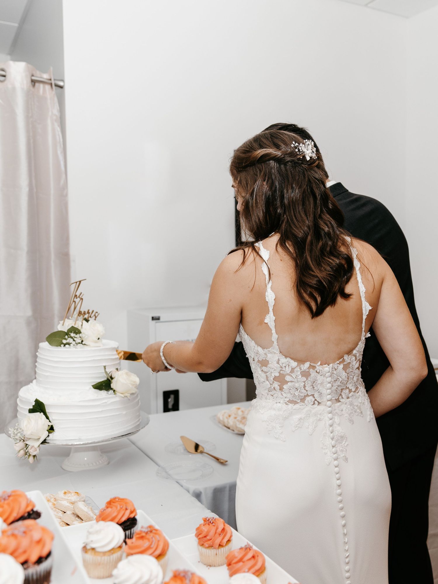 White wedding cake with floral decorations and cupcakes displayed at reception.