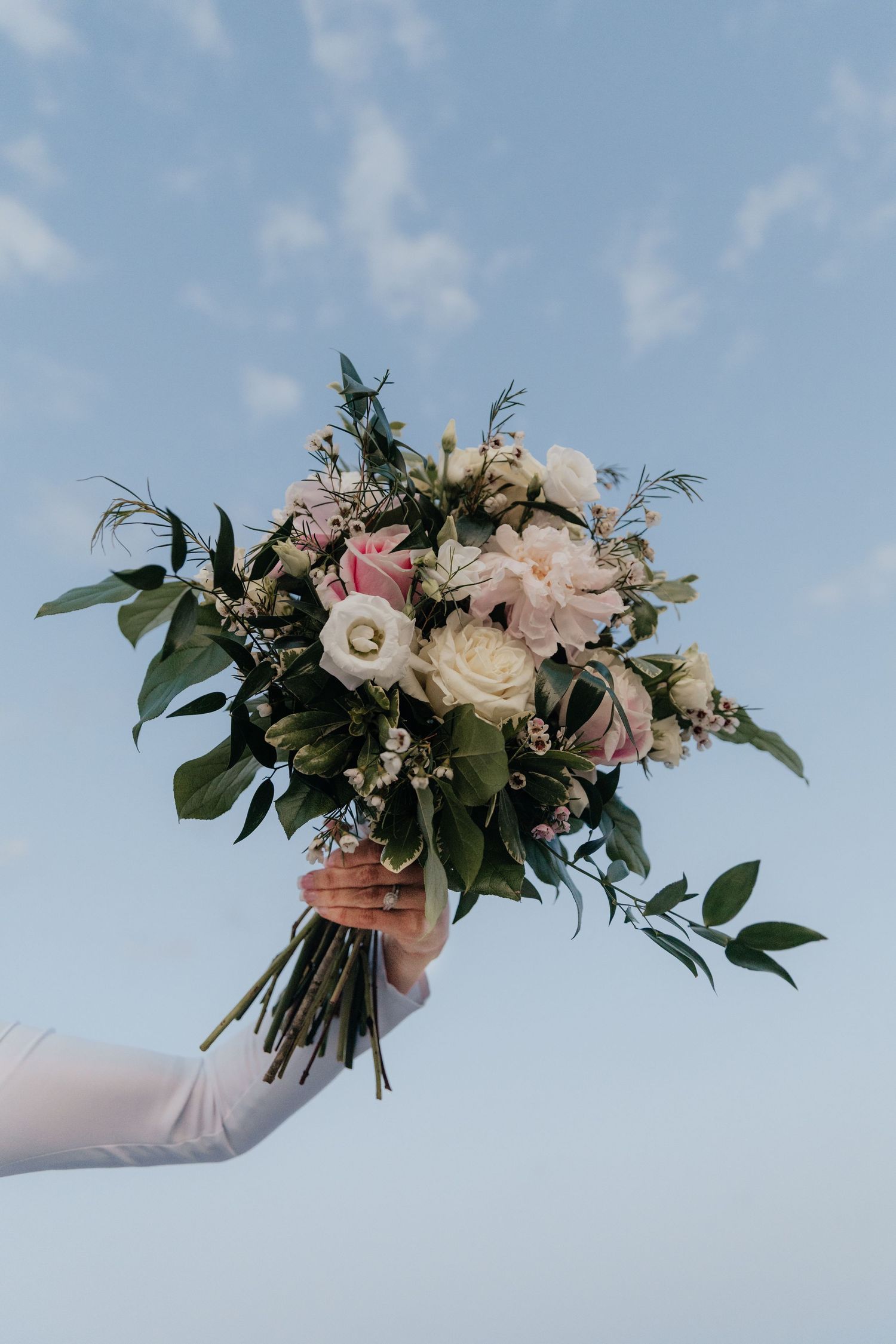 A hand holding an elegant wedding bouquet with roses and greenery against a blue sky.