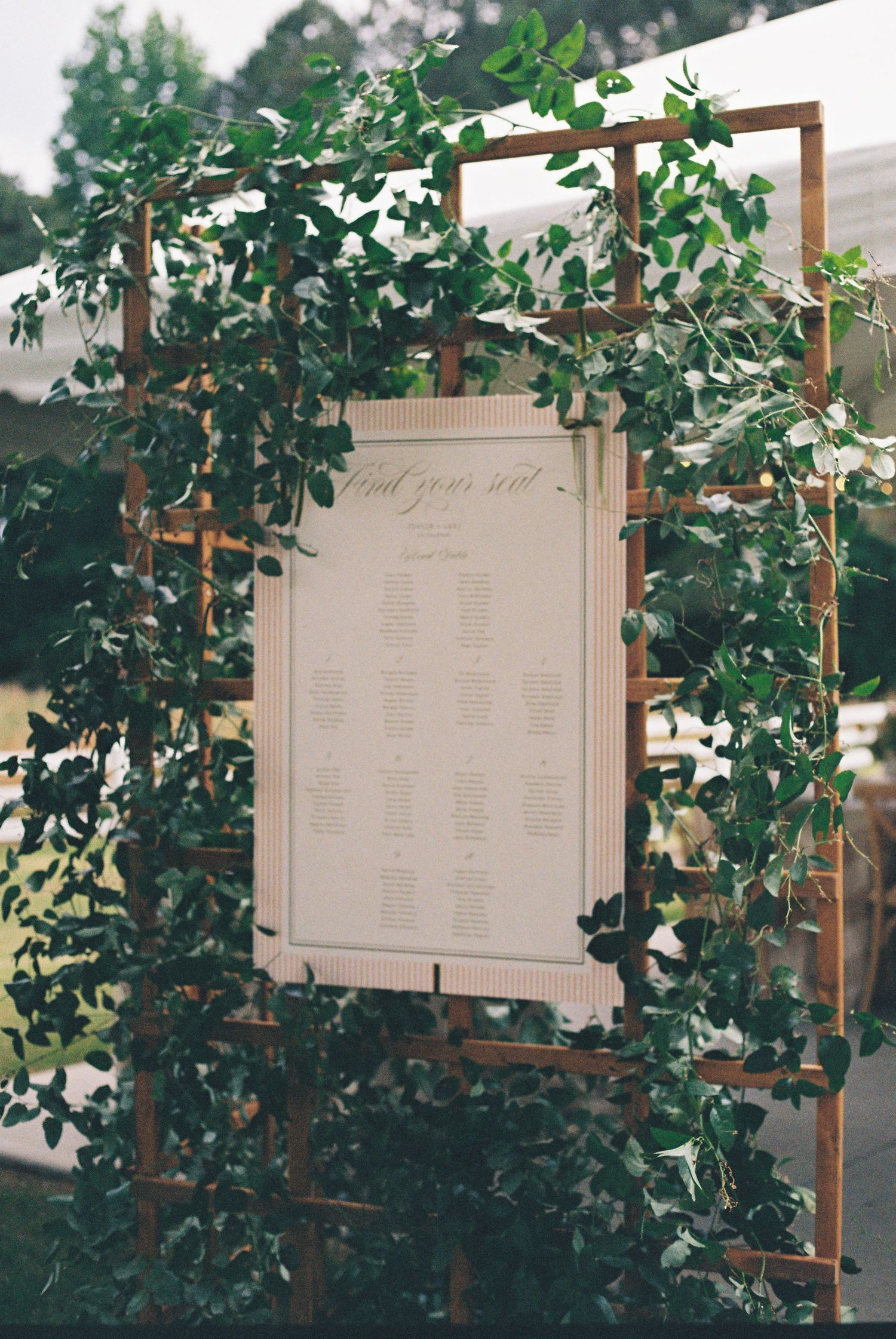 Wooden trellis arch covered in lush green ivy displaying an elegant wedding seating chart sign.