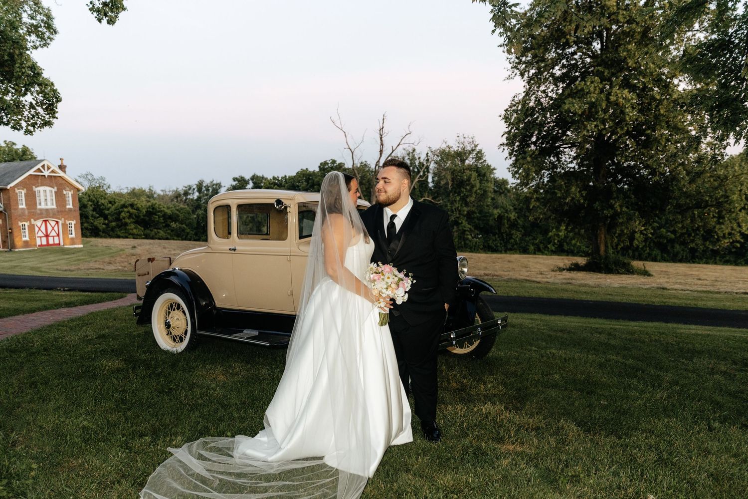 Wedding couple poses by vintage cream-colored car during outdoor sunset ceremony.