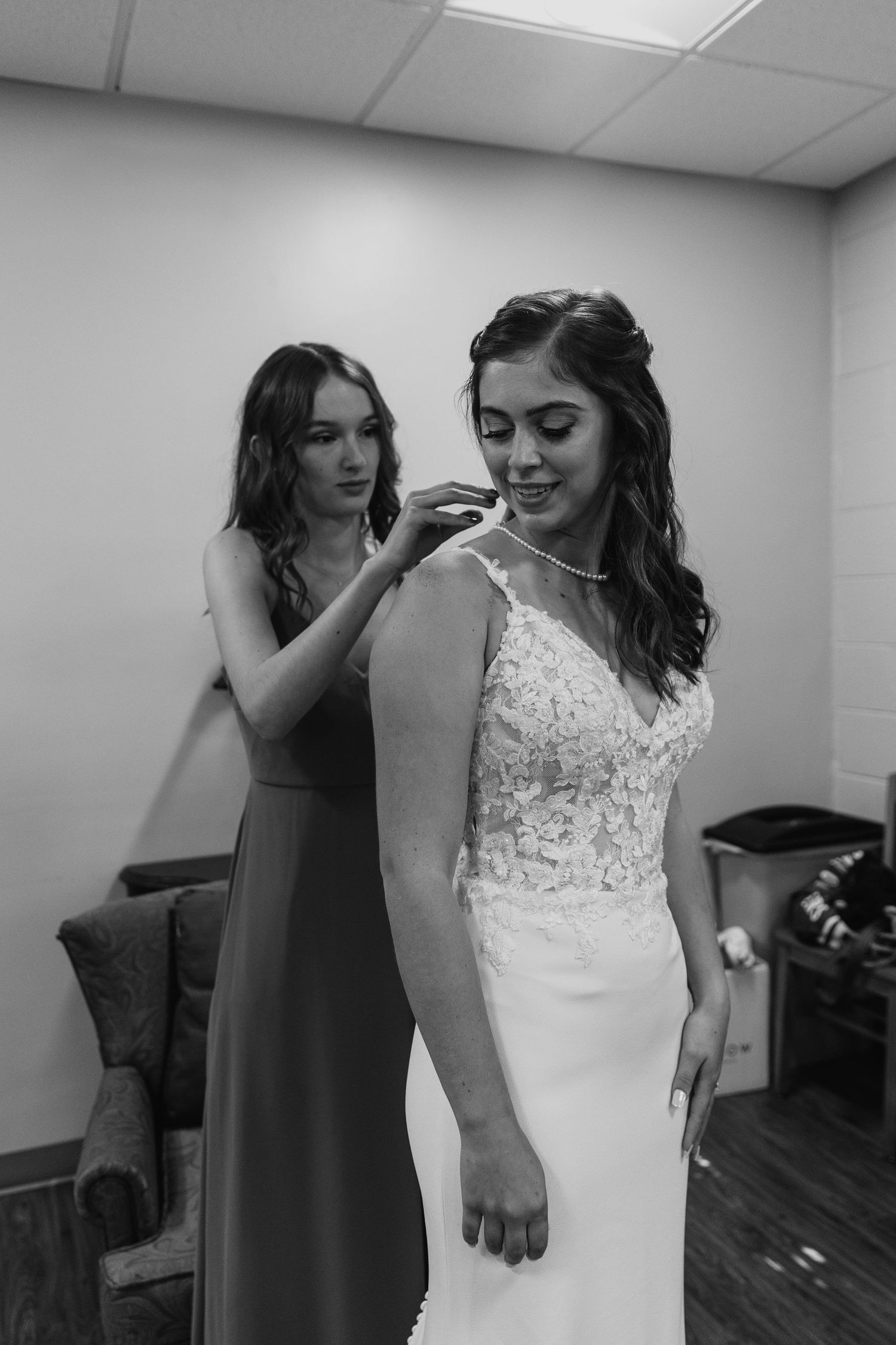 A black and white photo of a bridesmaid helping the bride into her necklace during the getting ready.