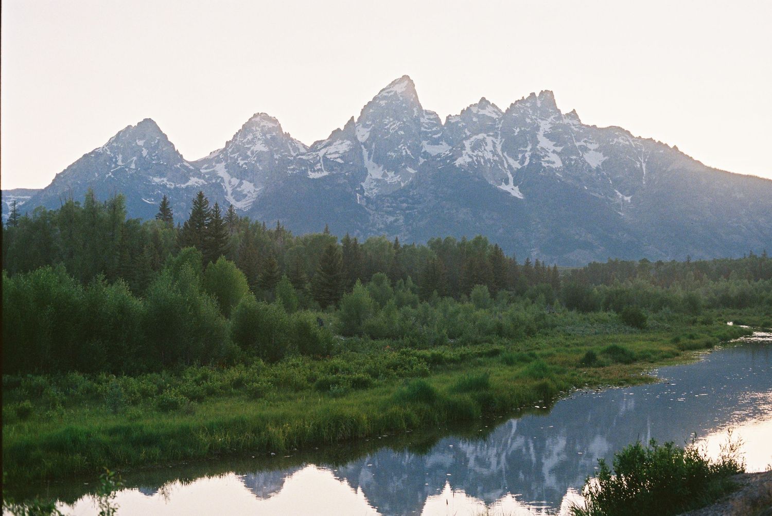 Snow-capped Grand Teton peaks reflected in calm river waters, surrounded by lush green forest at sunrise.