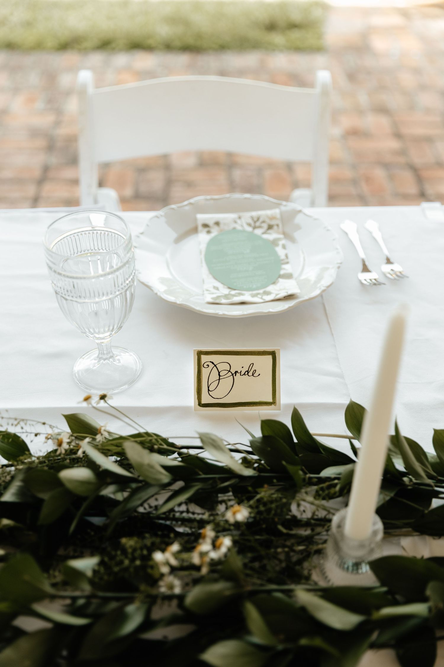Wedding table setting with crystal glass, white plate, and greenery garland centerpiece.