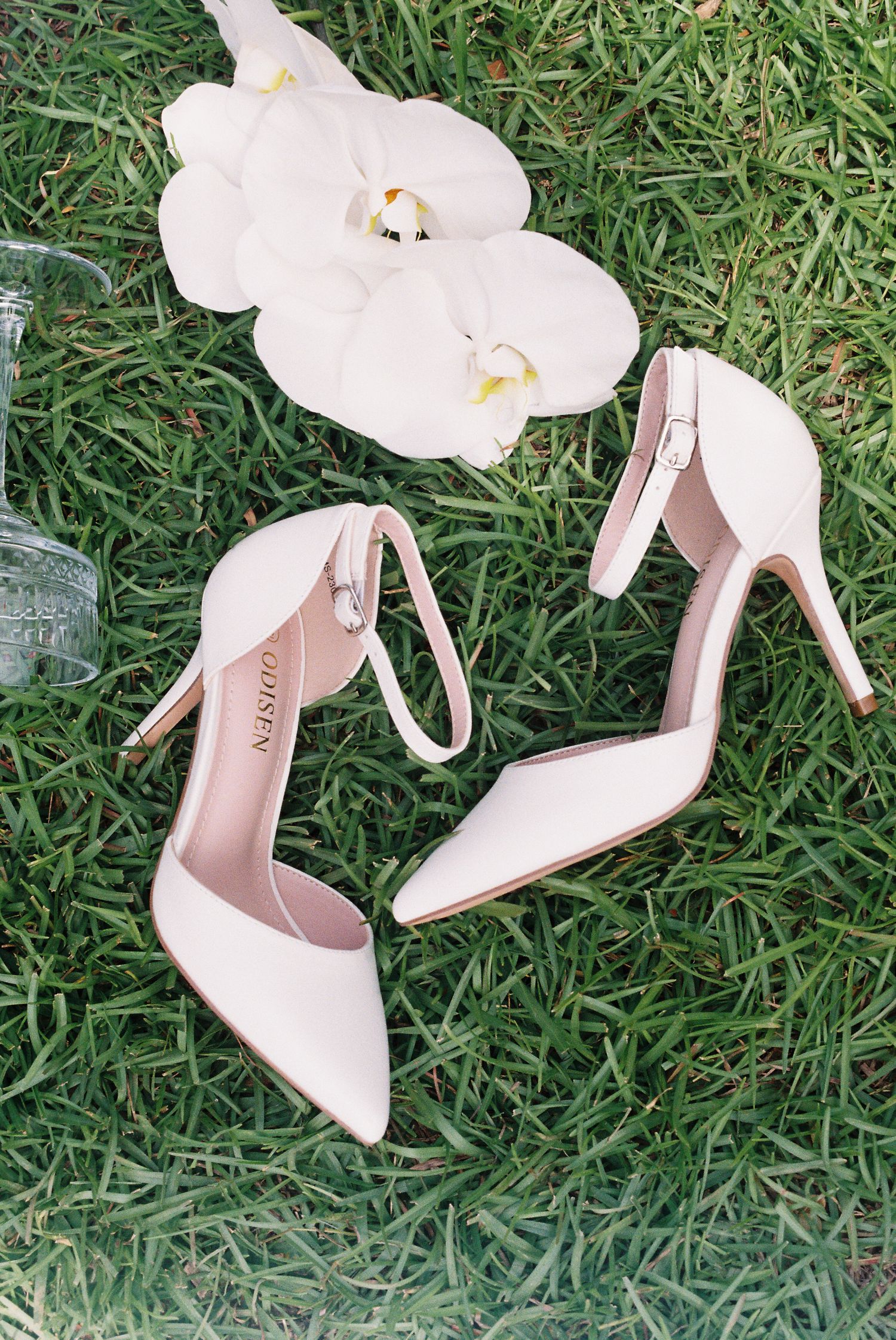 Elegant white bridal heels with floral details photographed on green grass.