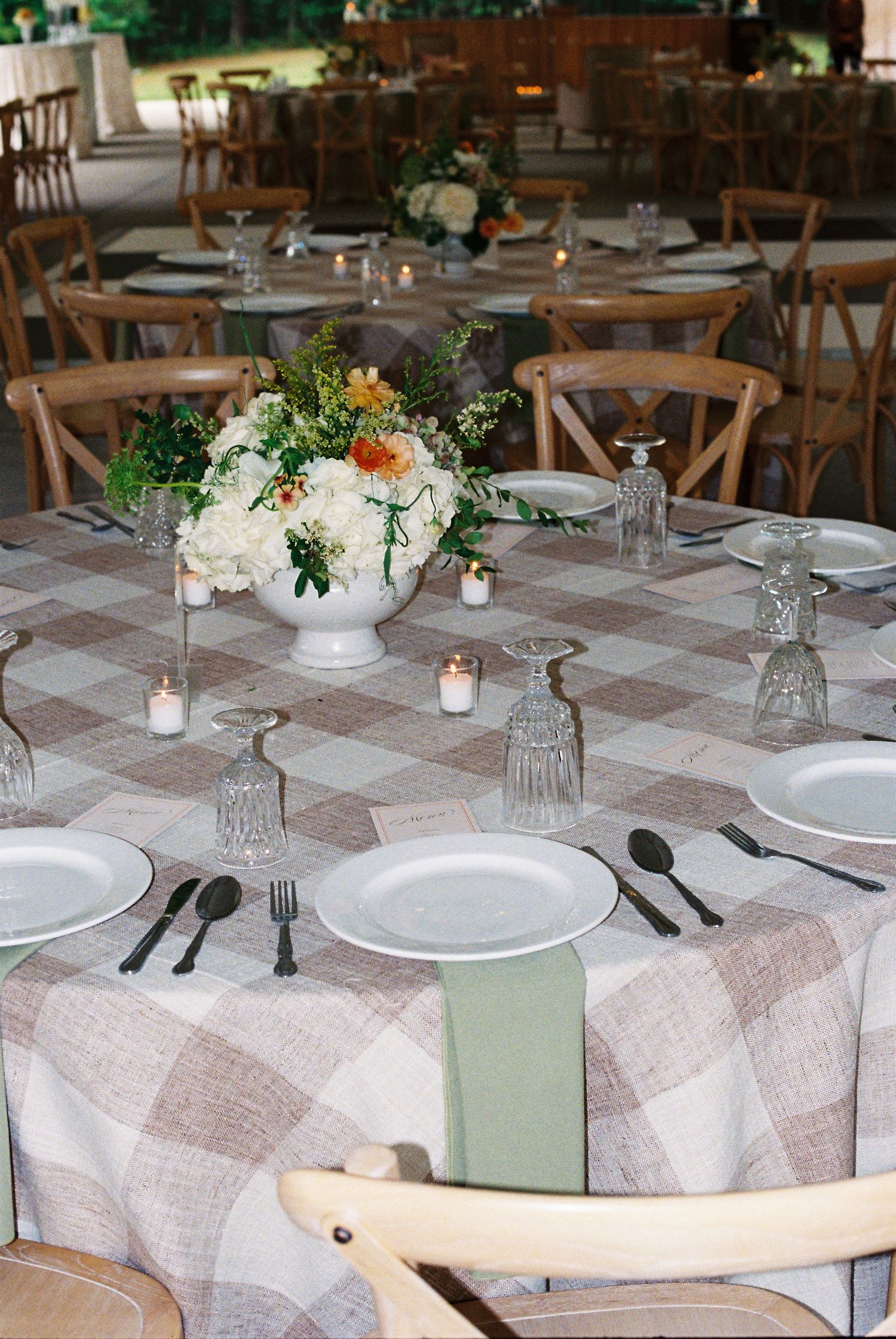 Elegant wedding reception tables with plaid linen, white floral centerpiece, sage napkins, crystal glasses, and black cutlery.