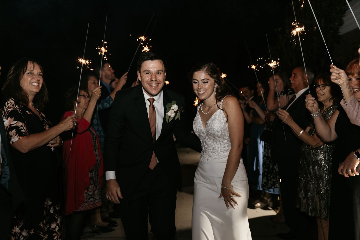 Newlyweds walk through sparkler tunnel surrounded by celebrating guests in formal attire.