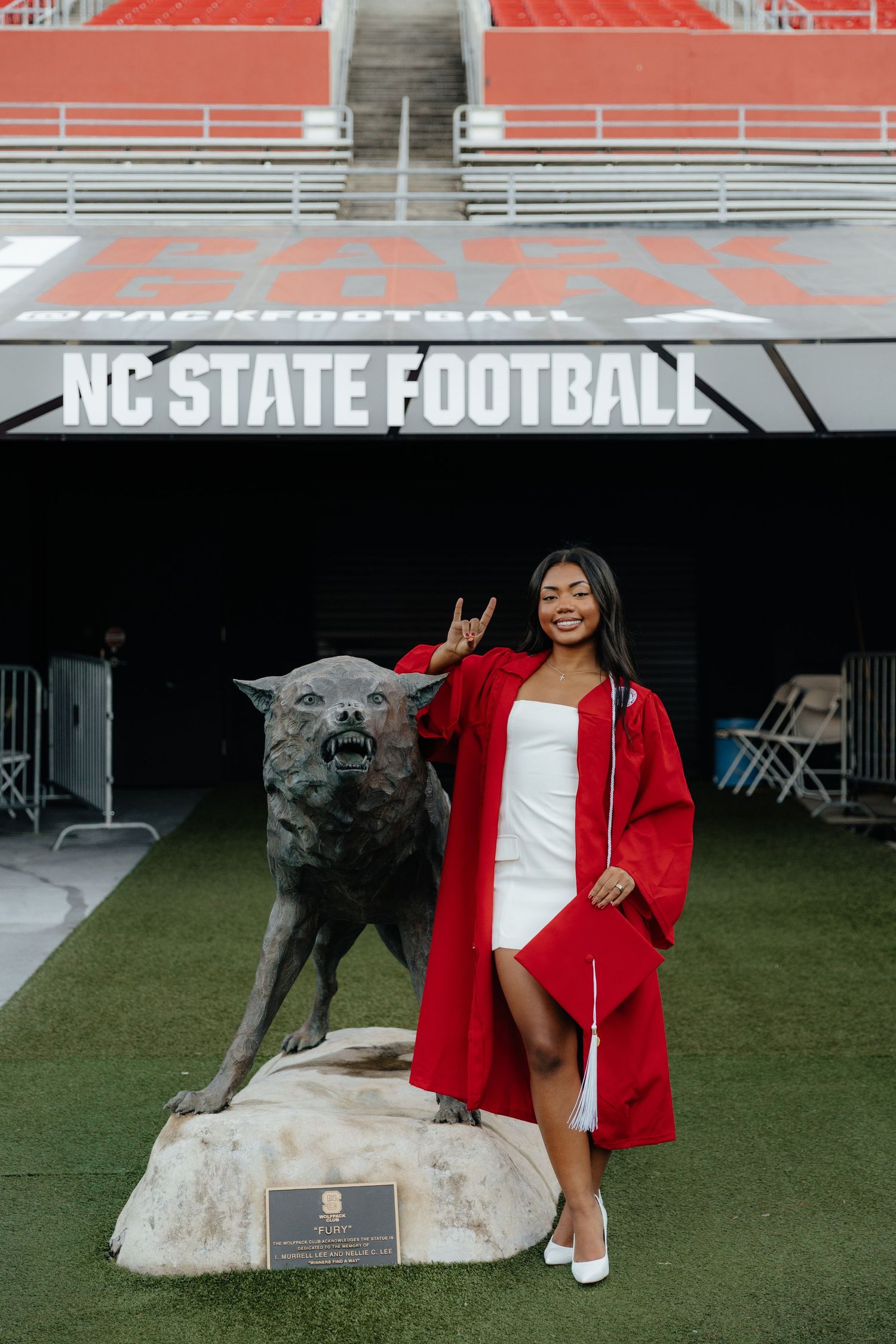 NC State graduate in red cap and gown poses with wolf statue at Carter-Finley Stadium football field.