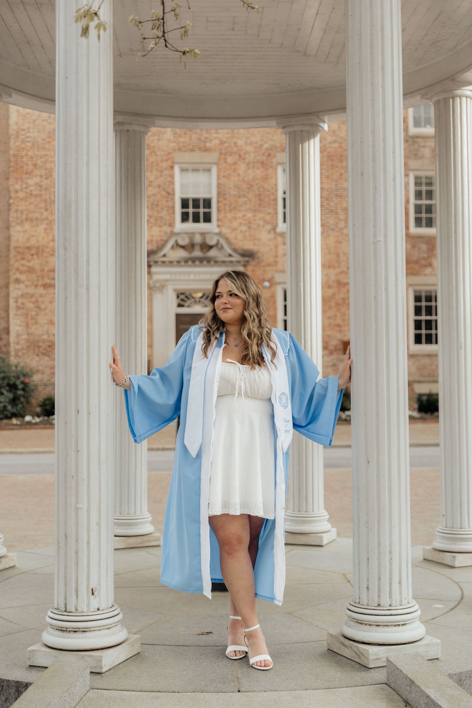 UNC graduate in light blue gown poses between white columns at Chapel Hill campus rotunda.