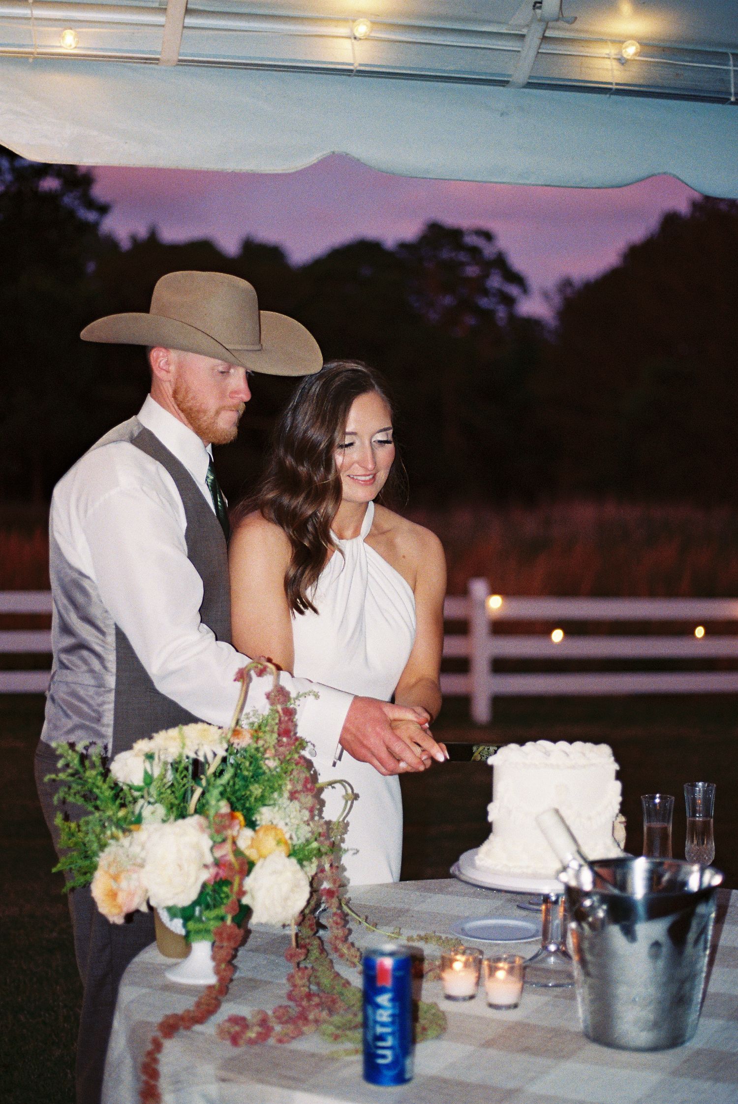 Bride and groom cutting wedding cake outdoors at sunset, groom wearing cowboy hat and vest at rustic country wedding reception.