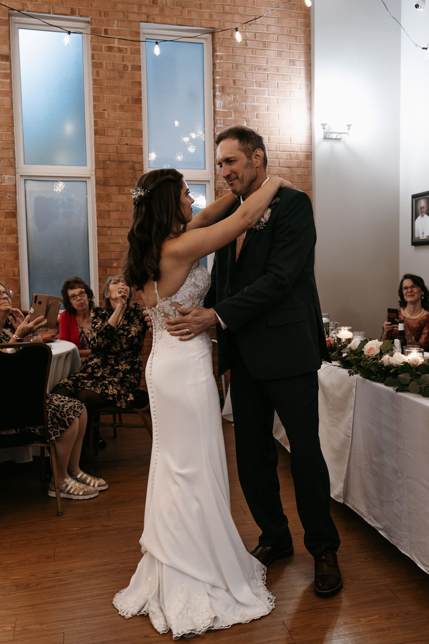 Couples slow dancing on a wooden dance floor during a wedding reception with exposed brick walls.