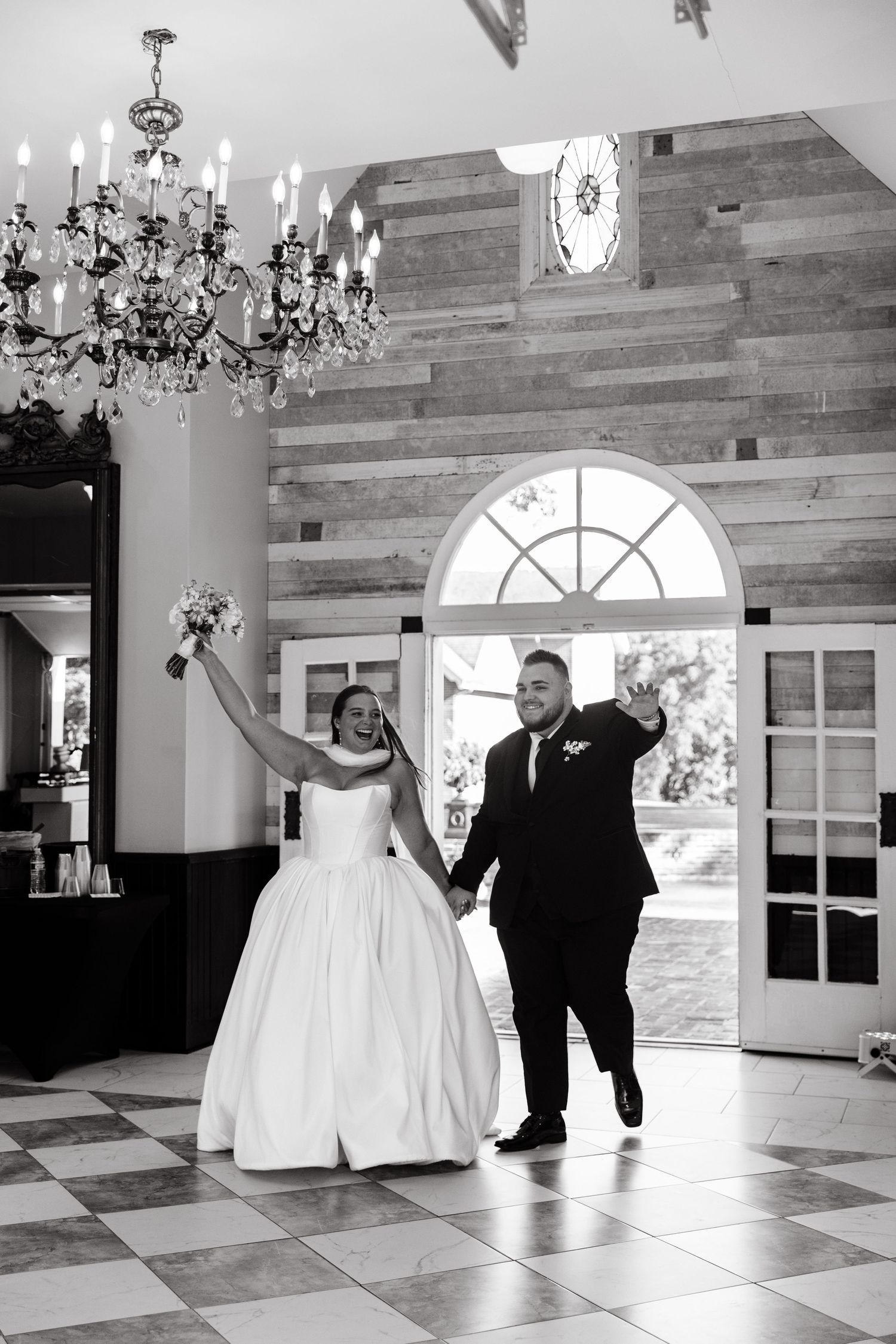 Black and white photo of newlyweds dancing in an elegant ballroom with chandeliers.