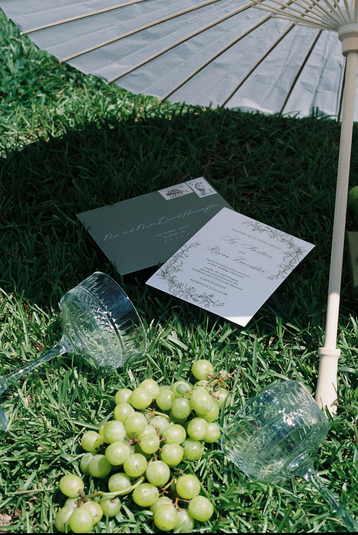 Wedding invitations and green grapes arranged on grass under a decorative umbrella.